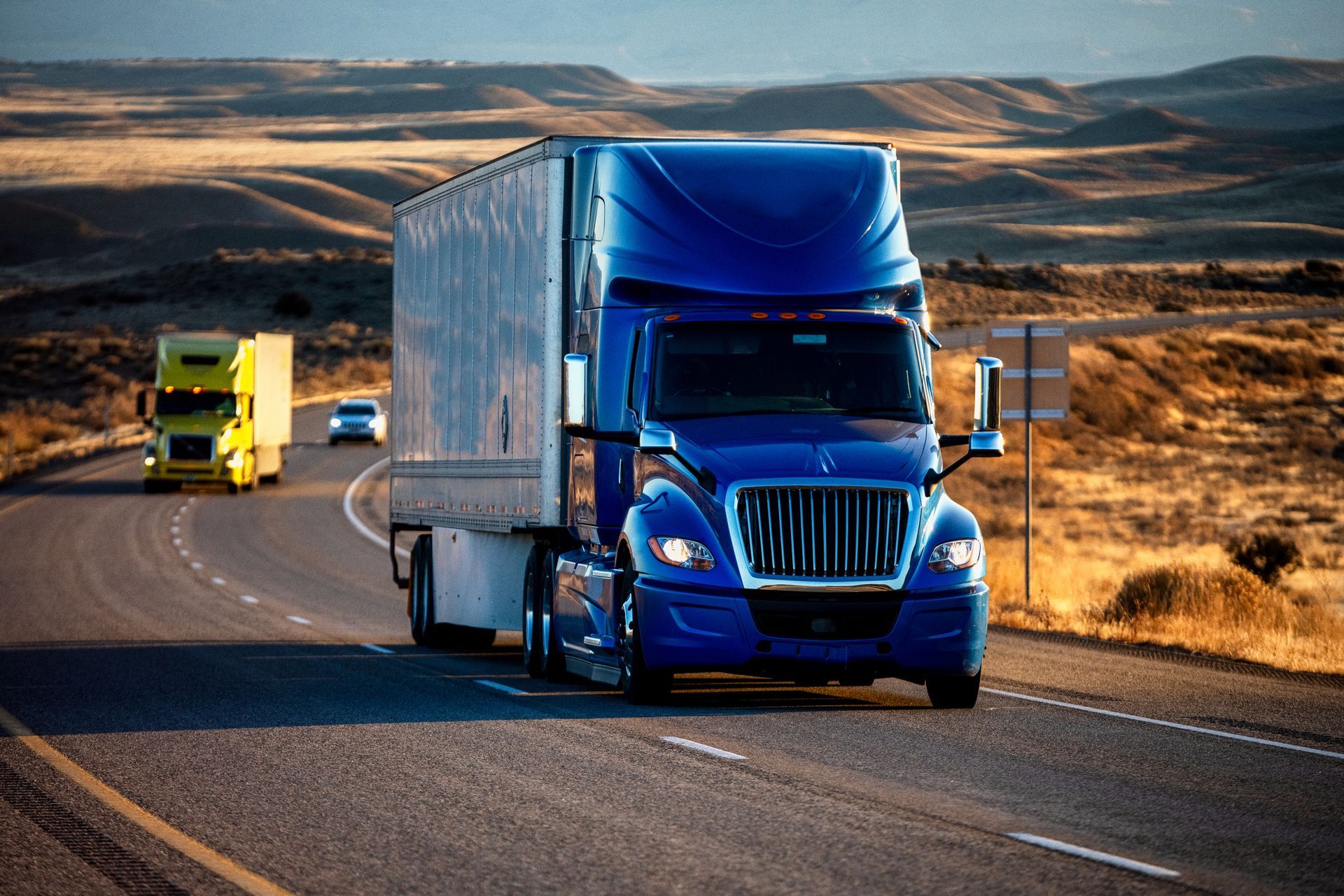Blue semi-truck driving on a highway with two other vehicles in a desert setting.