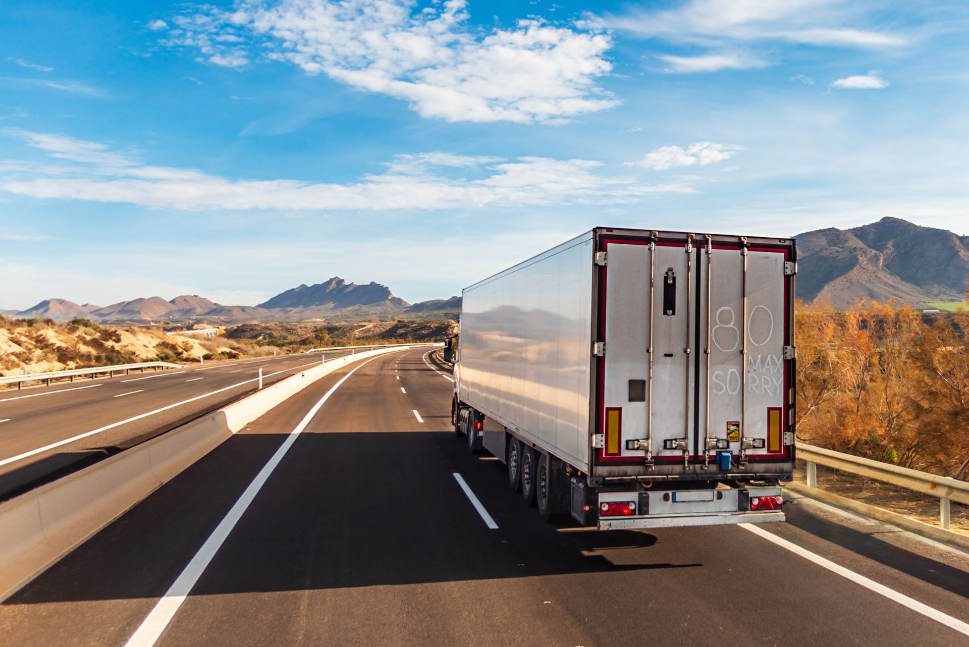 A semi-truck on a highway, driving towards mountains under a blue sky.