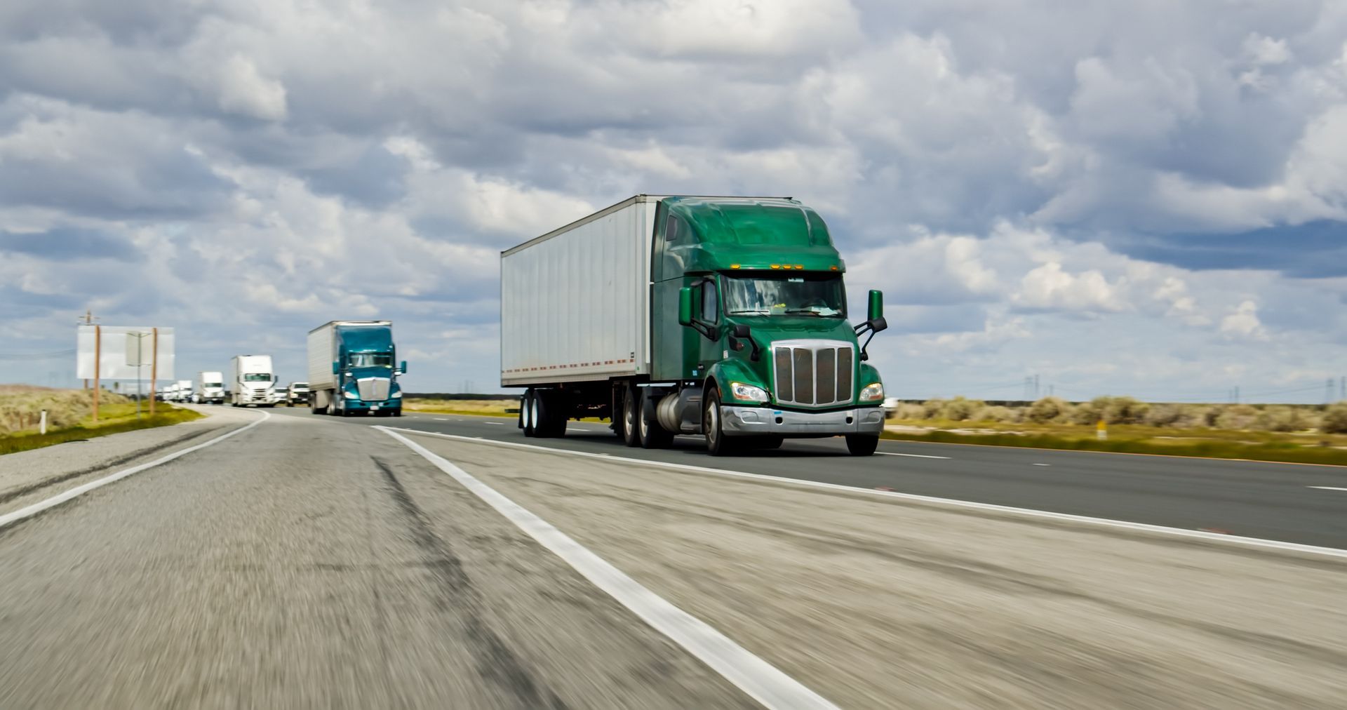 Green semi-truck driving on highway, with other trucks in the distance, under a cloudy sky.