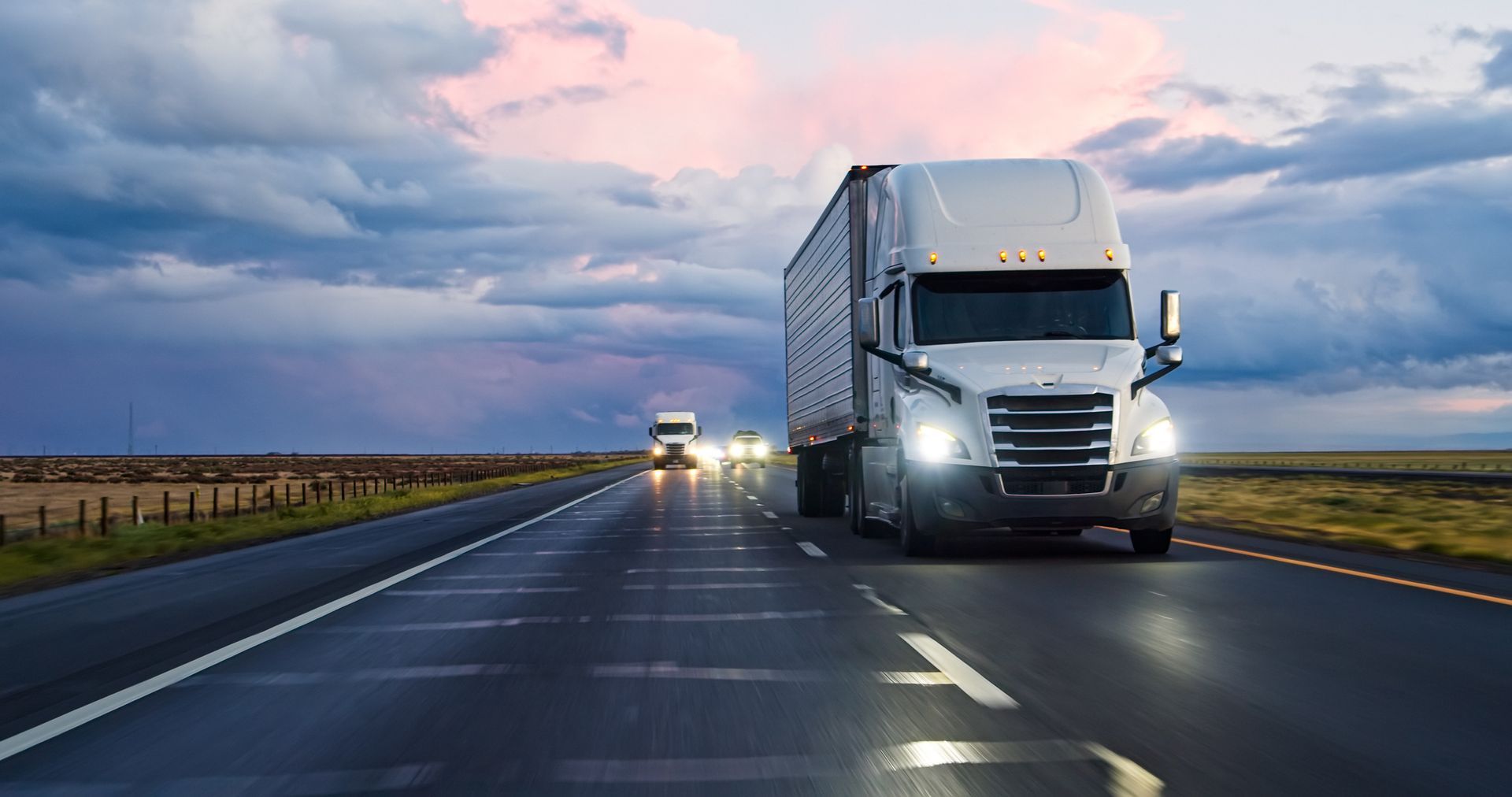 Two semi-trucks driving on a wet highway under a cloudy sky at dusk.