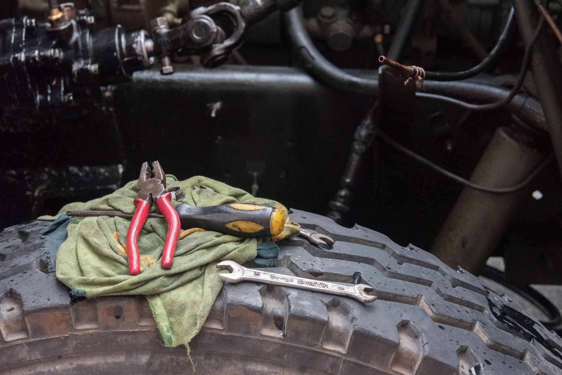 Tools (pliers, wrench, screwdriver) on a rag atop a truck tire, near engine components.