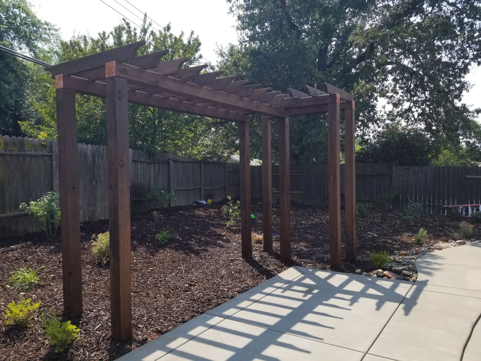 A wooden pergola in a backyard with a fence in the background