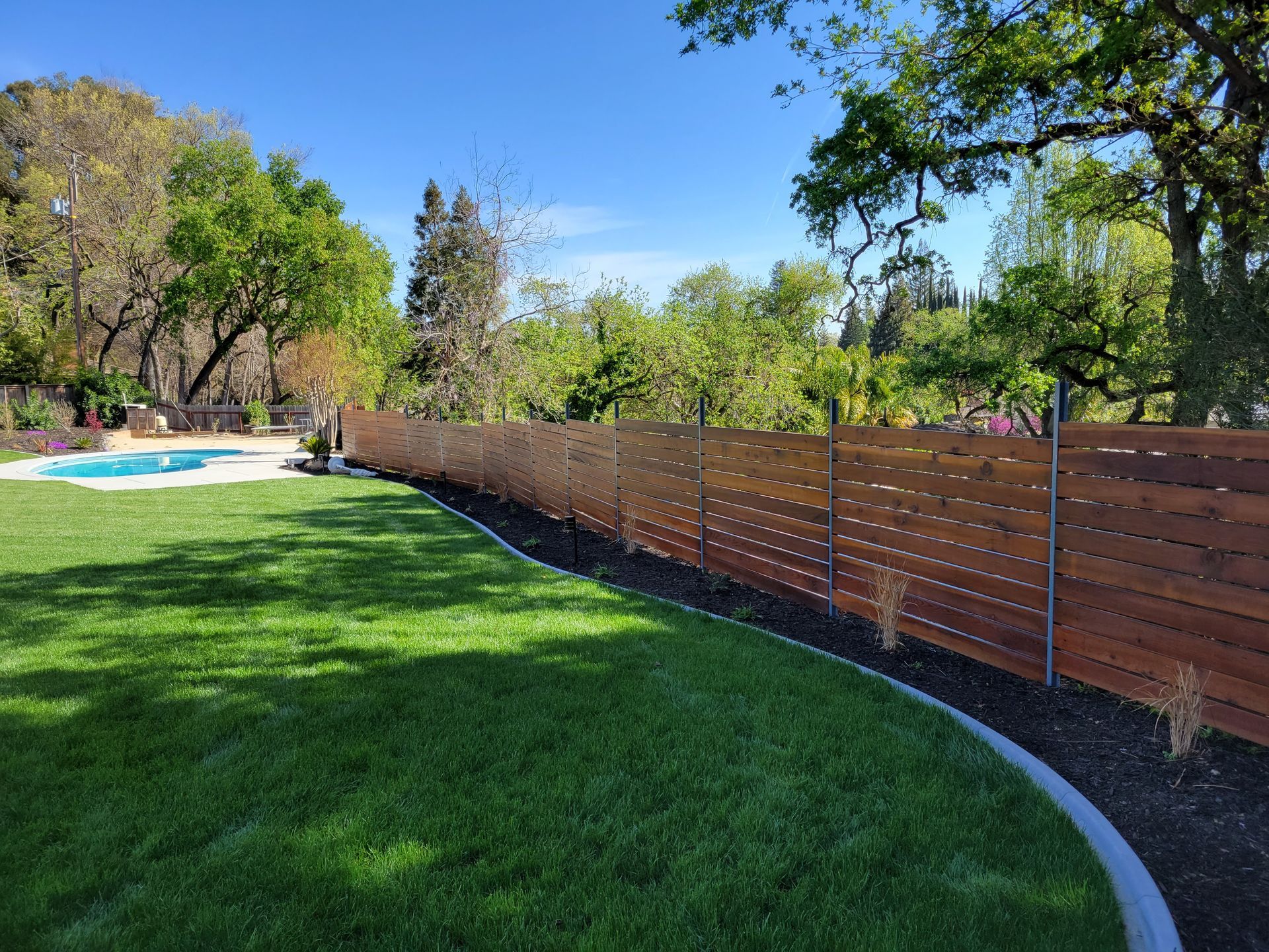 A wooden fence surrounds a lush green yard with a pool in the background.