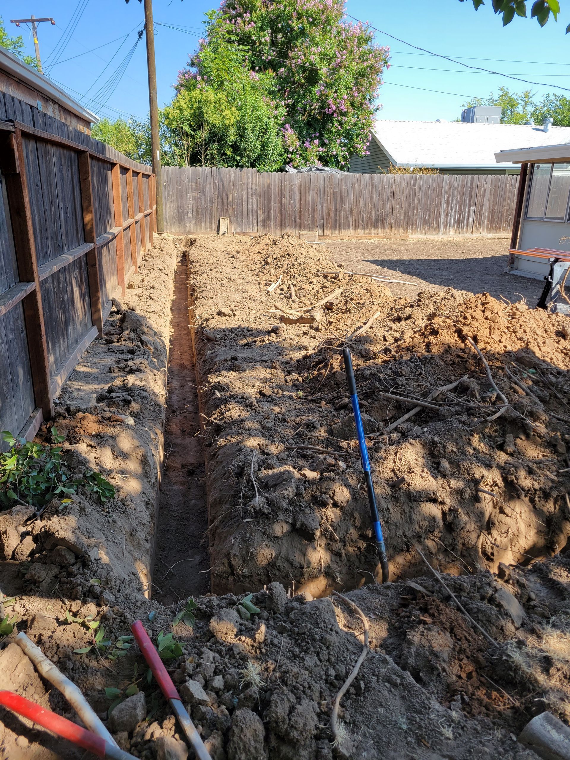 A shovel is sitting in the dirt in a backyard next to a wooden fence.