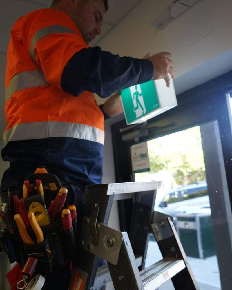 A Man Is Standing On A Ladder Holding A Green Exit Sign — Firewerx In Bathurst, NSW