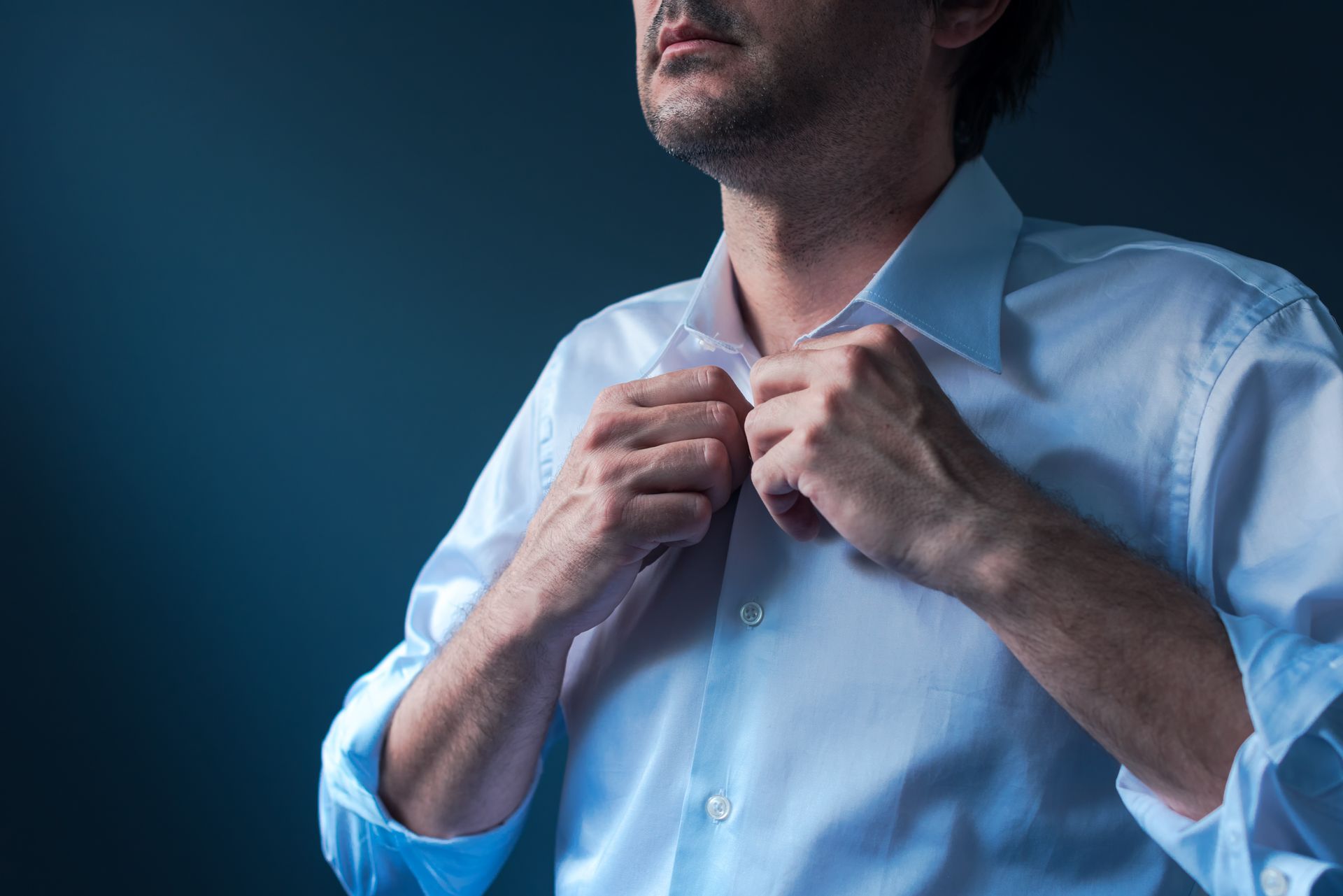 Man buttoning a white collared shirt against a dark background.