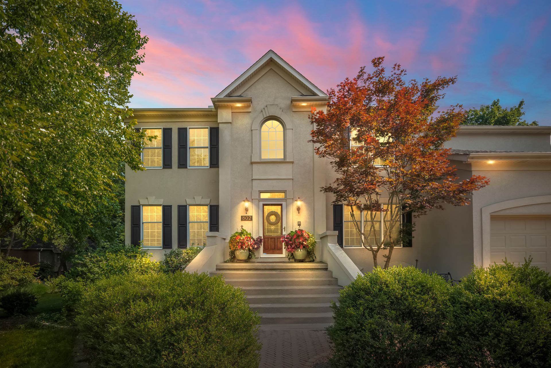 Two-story beige house with steps, plants, and a colorful sunset sky.