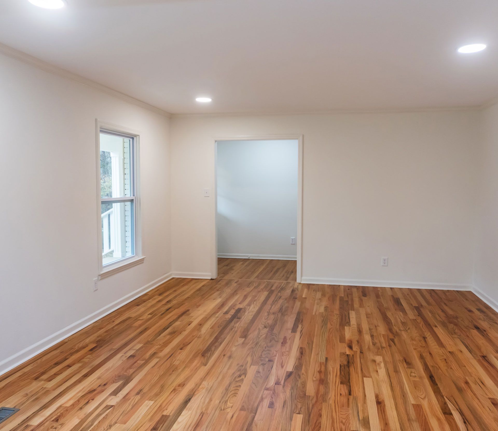Empty room with hardwood floors, white walls, and a doorway. Sunlight streams in from a window.