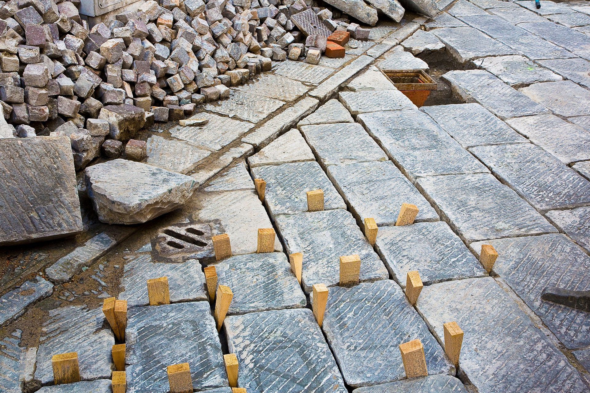 Construction site: paving stones being laid with wooden spacers. Piles of stone and debris.