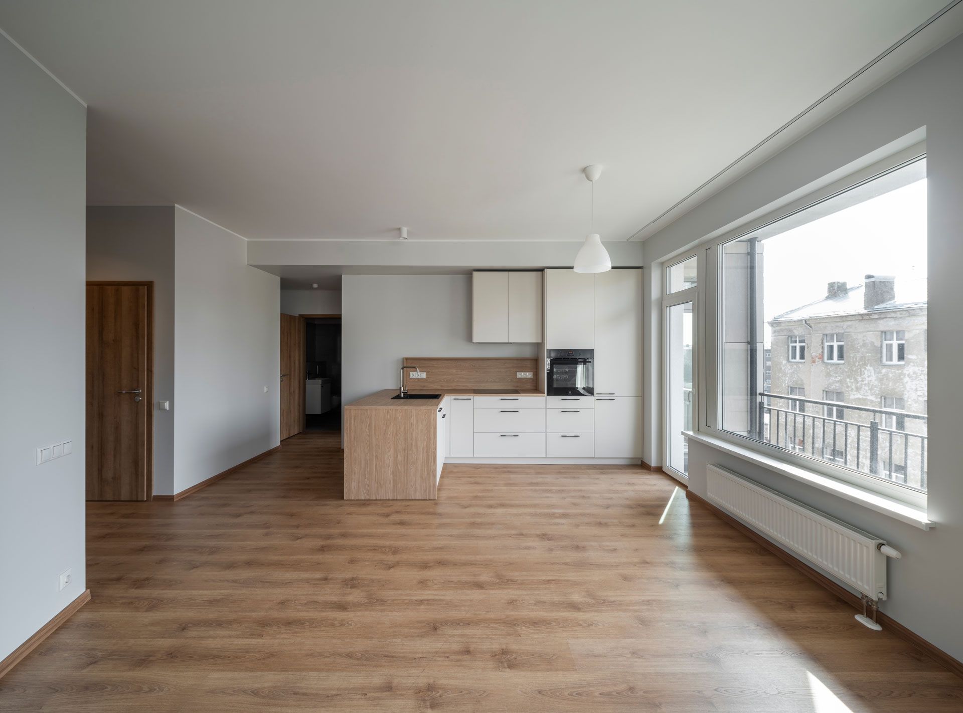 Empty open-plan apartment with wood floors, white cabinets, and large windows.