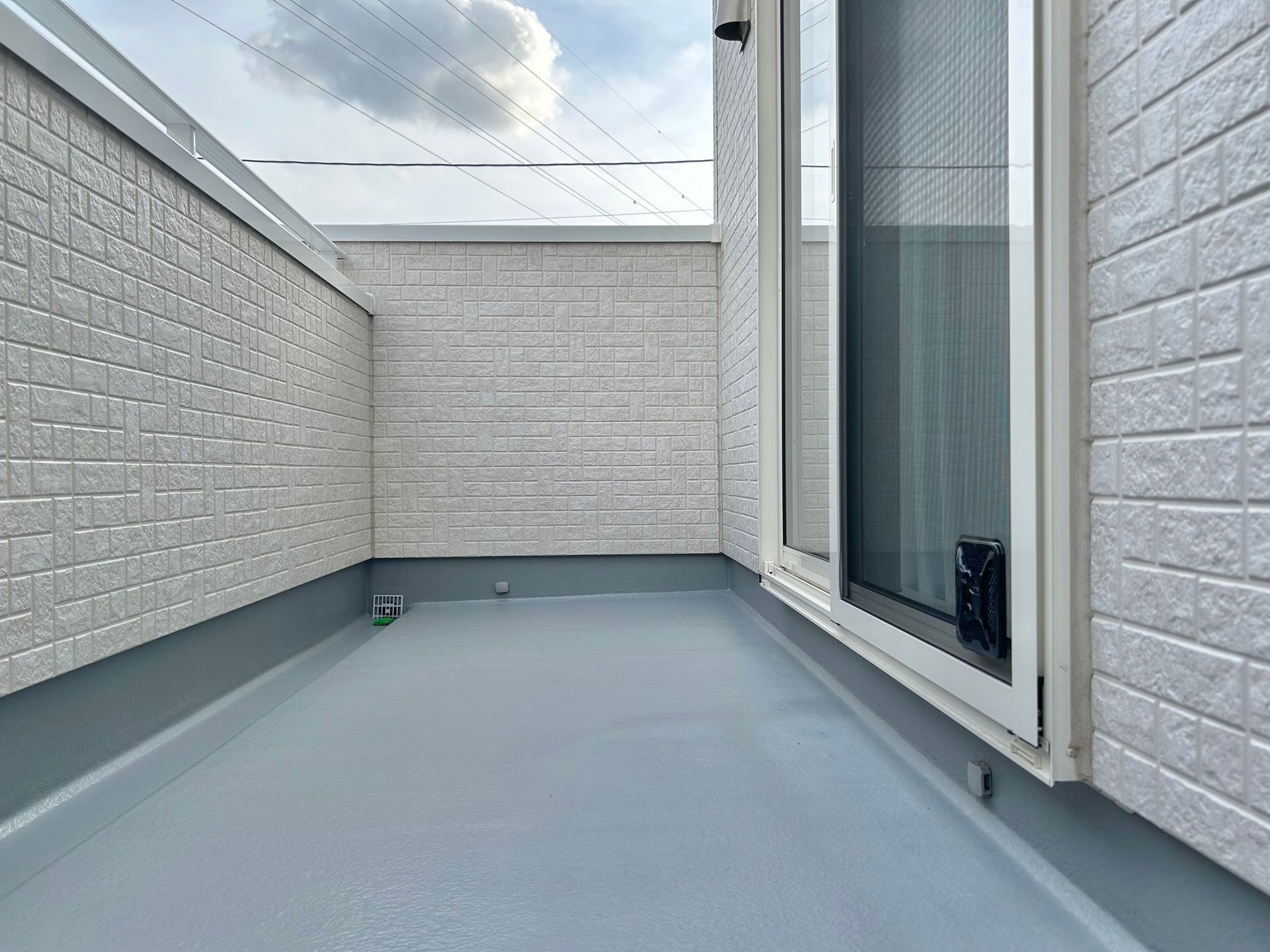 Small, enclosed balcony with light gray floor and brick-patterned walls; a glass door on the right.