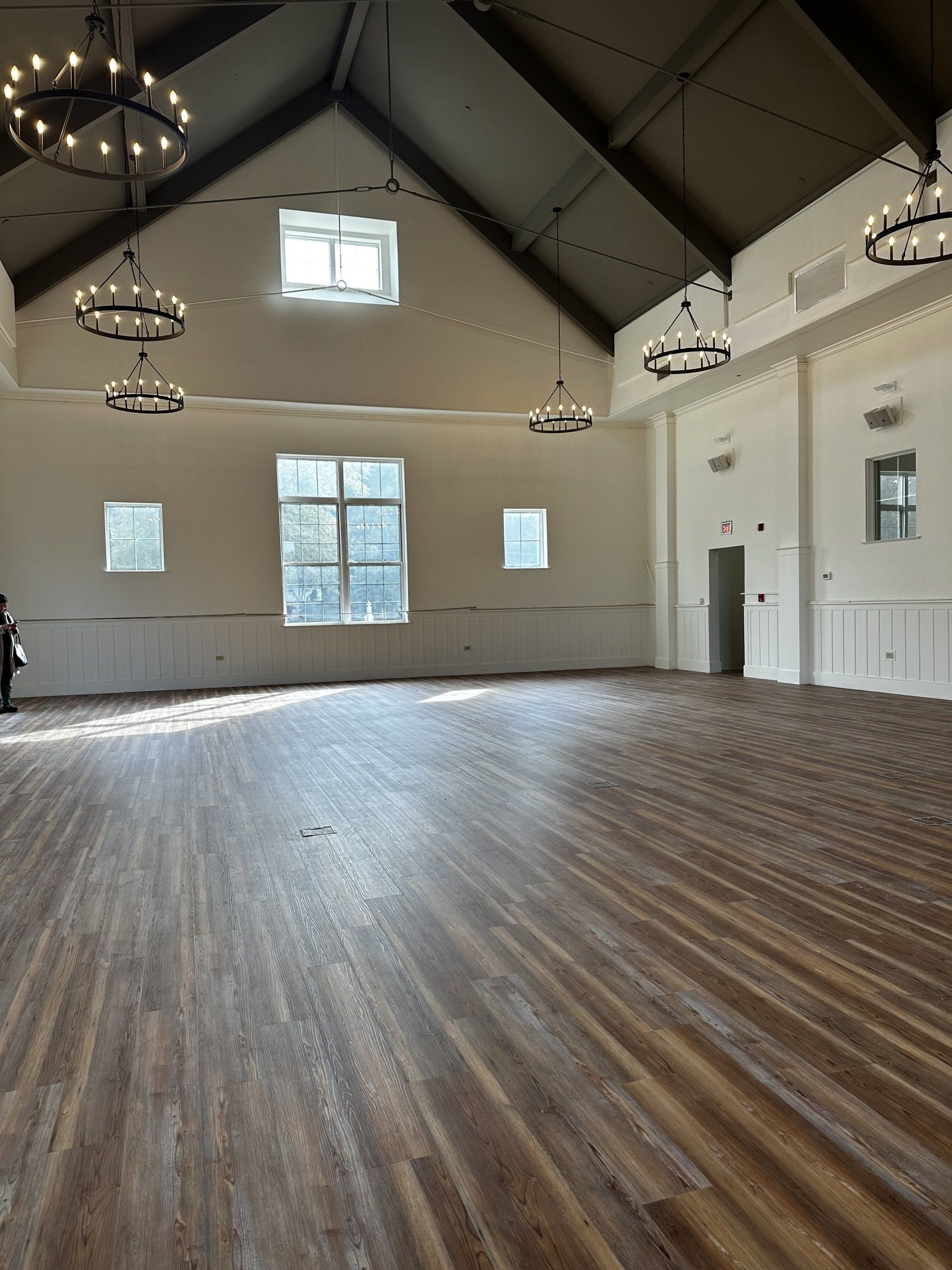 Empty ballroom with wood floor, white walls, and chandeliers. Sunlight streams in from windows.