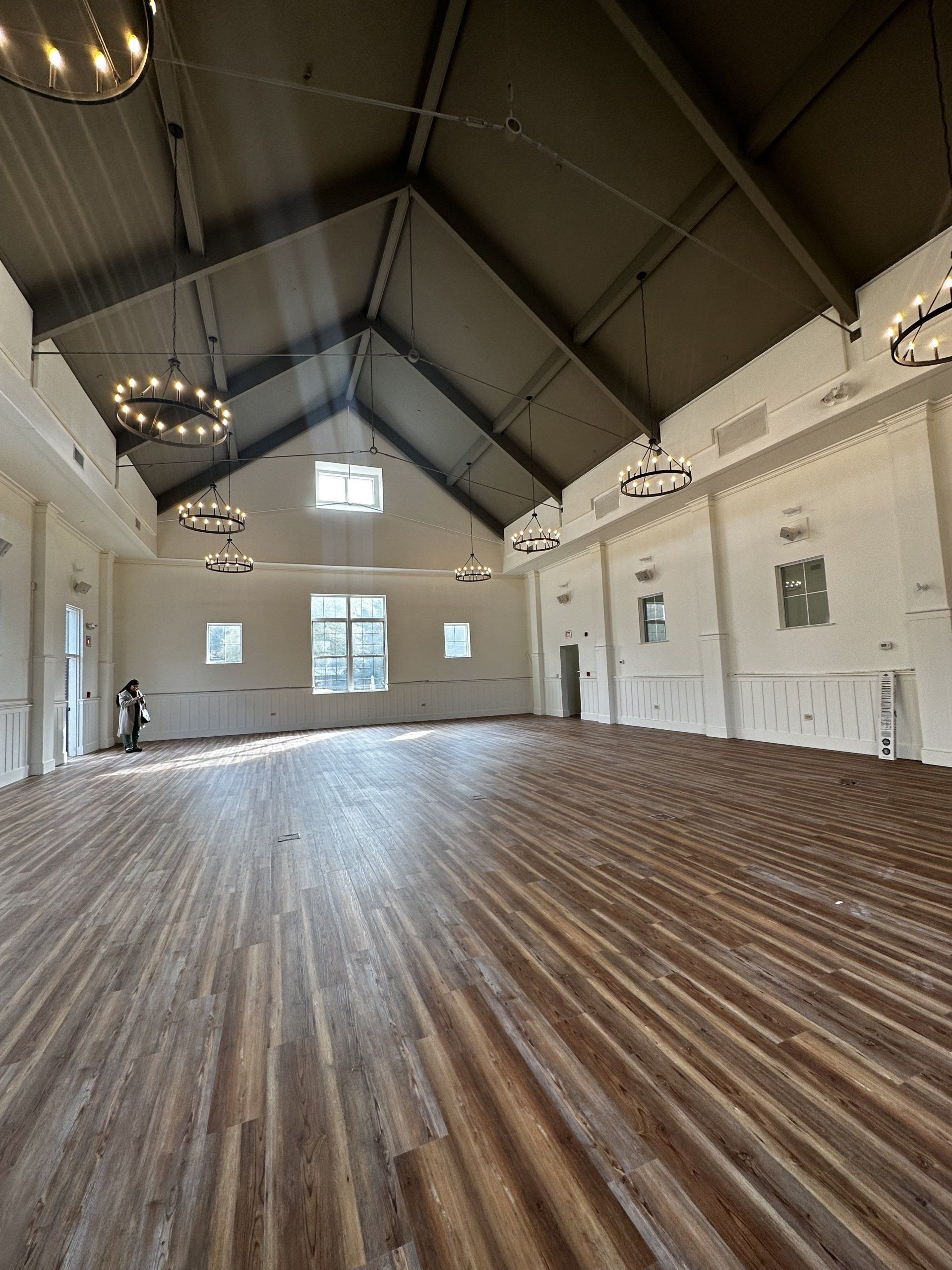 Spacious event hall with wood floor, white walls, dark ceiling, and round chandeliers.