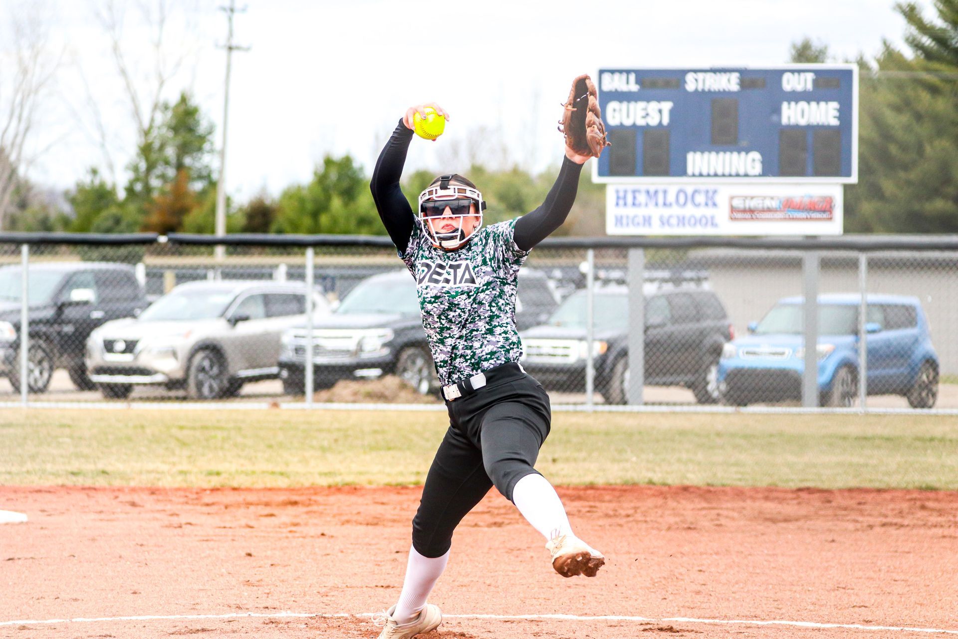 A softball player is throwing a ball in front of a scoreboard.