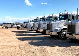 A row of semi trucks are parked in a dirt lot.