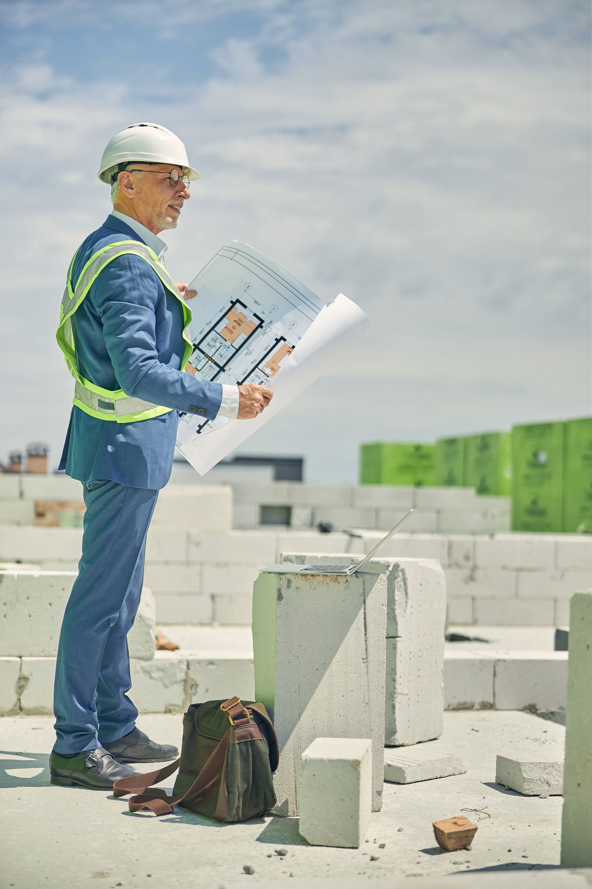 A man in a suit and hard hat is standing on a construction site holding a blueprint.