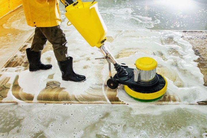 Worker pressure-washing a foam-covered floor with a yellow cleaning machine and boots in view