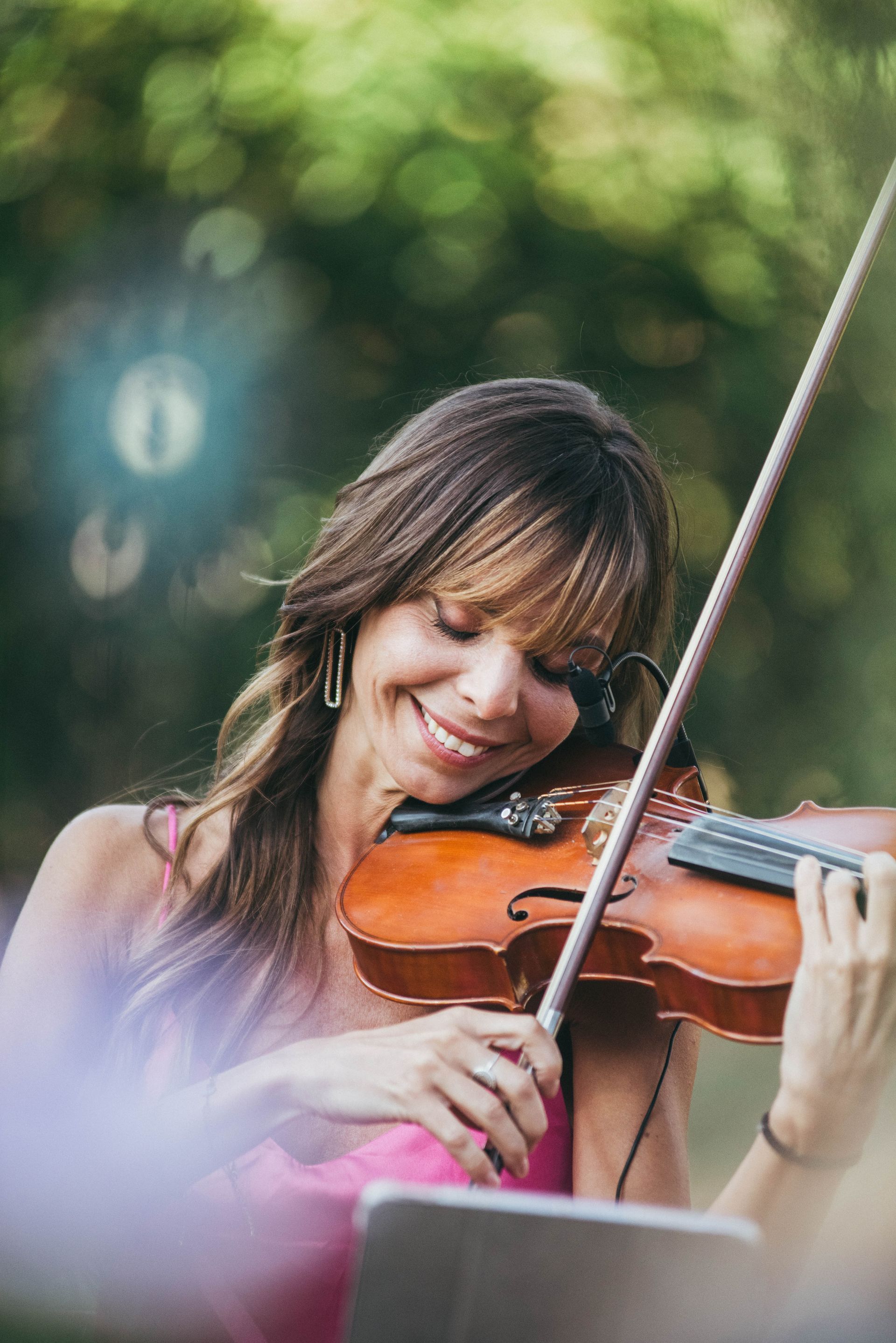 Donna che suona il violino all'aperto, sorridente, con un microfono in cuffia, sfondo verde sfocato.