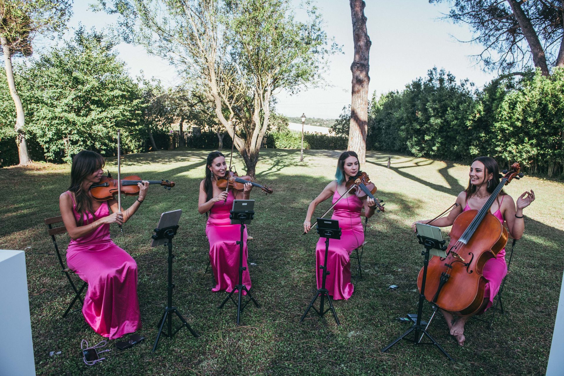 Quattro donne in abiti rosa suonano strumenti a corda all'aperto in una giornata di sole.