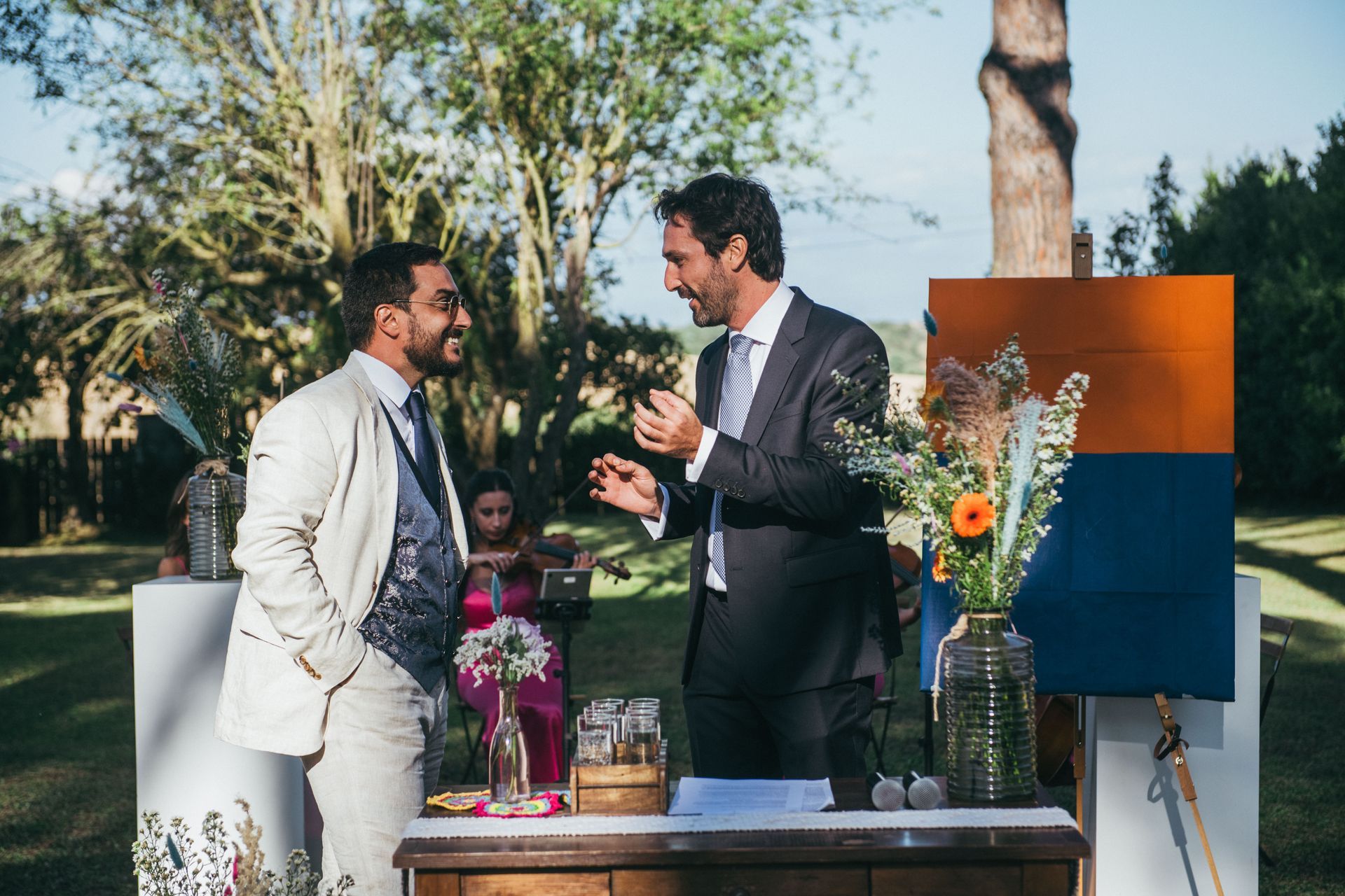 Due uomini in giacca e cravatta parlano durante un evento all'aperto, tra fiori e uno sfondo colorato.