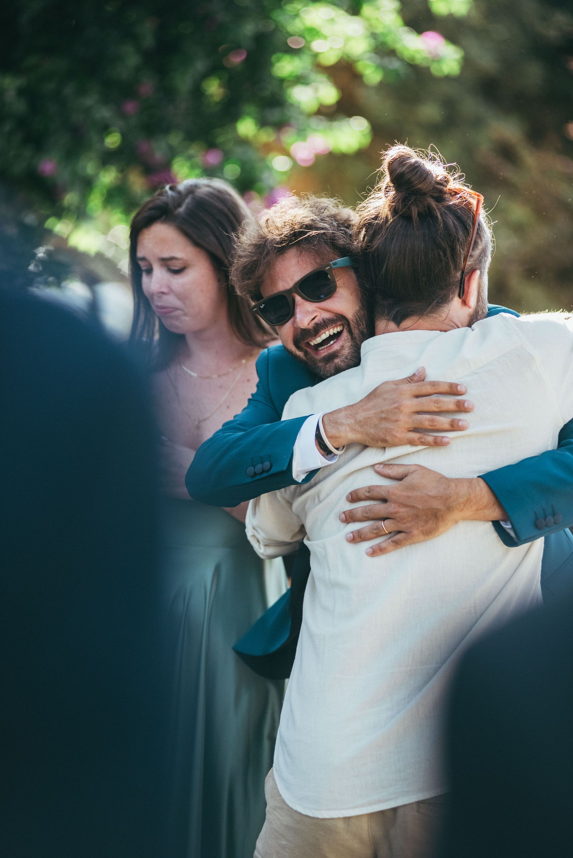 Un uomo con gli occhiali da sole abbraccia un altro uomo. Una donna con un vestito verde osserva. Scenario all'aperto.