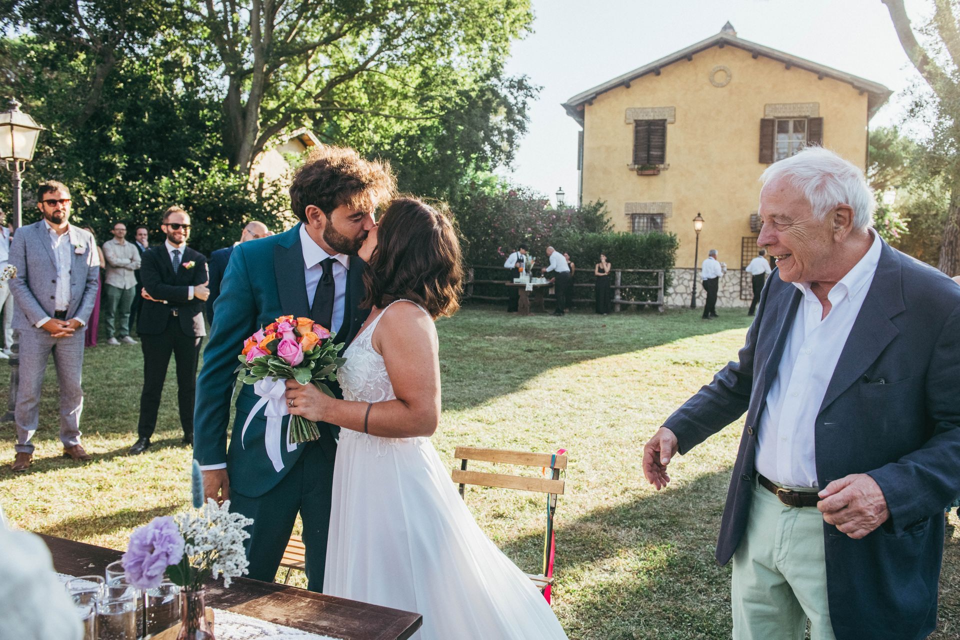 Coppia che si bacia durante una cerimonia nuziale all'aperto, uomo che si avvicina; edificio giallo sullo sfondo.