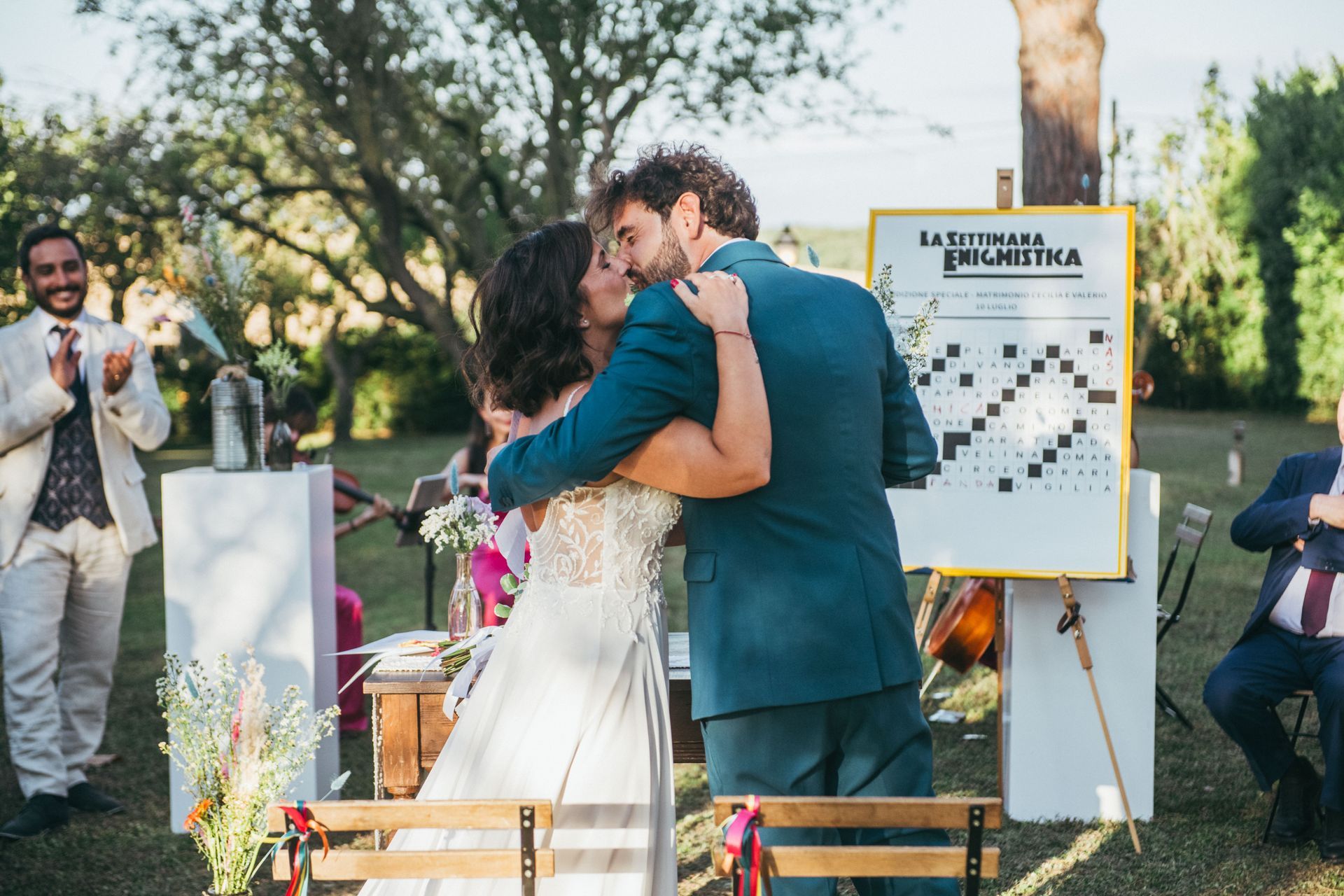 Coppia che si bacia a un matrimonio all'aperto; poster sul cavalletto; ospiti che applaudono; giornata di sole.