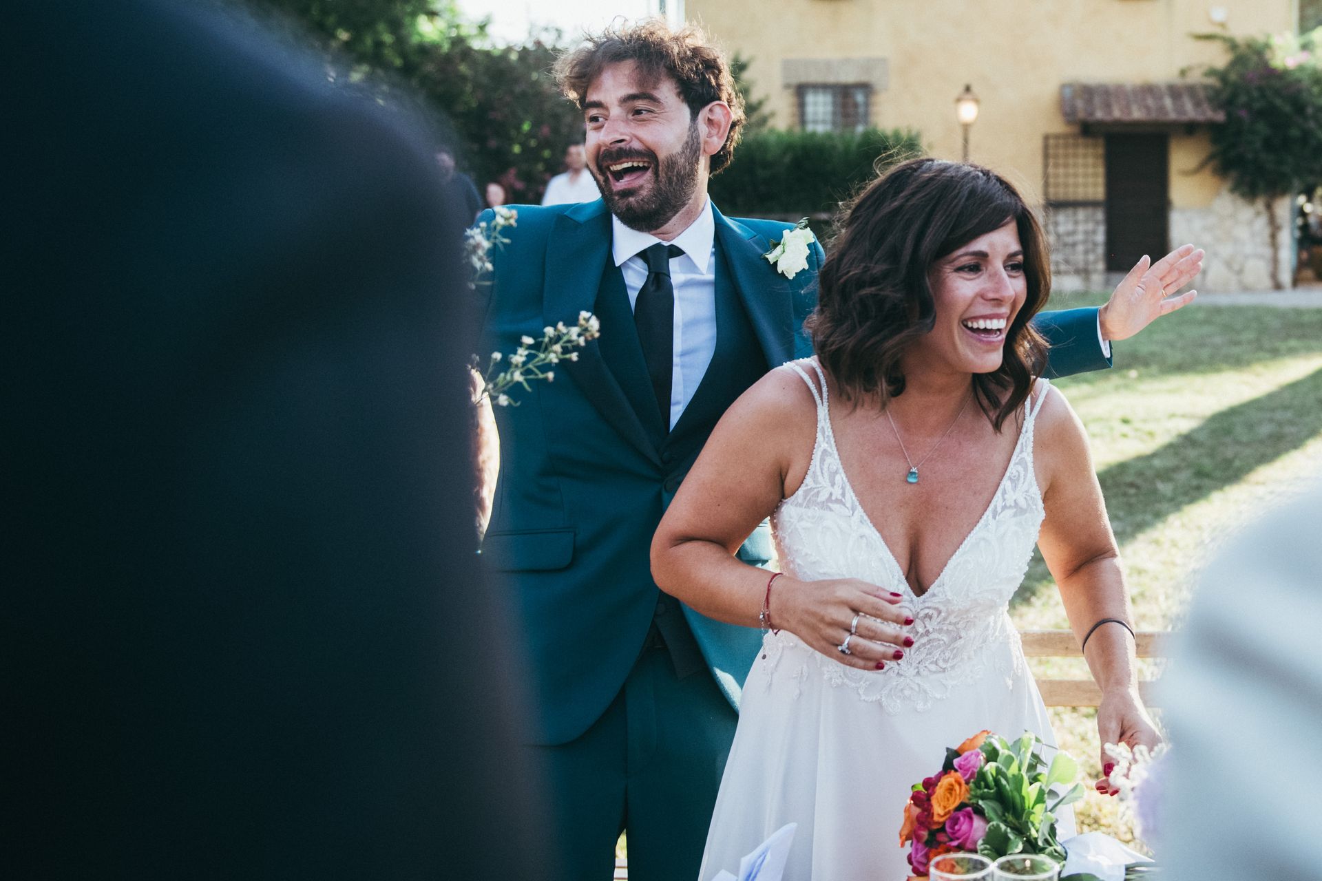Gli sposi ridono gioiosamente, festeggiando. Lo sposo in abito blu, la sposa in abito bianco, all'aperto.