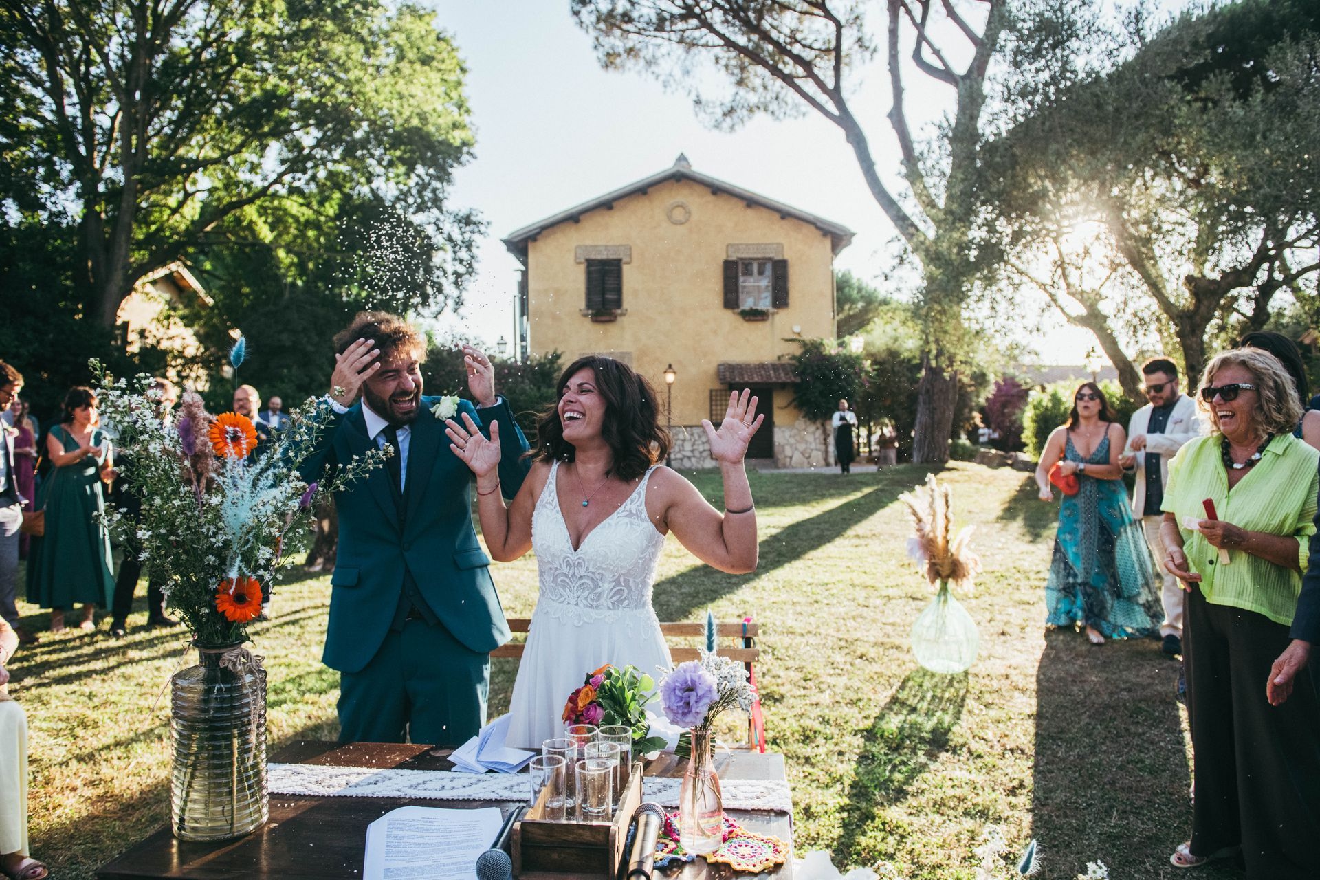 Gli sposi festeggiano in una cerimonia in un giardino soleggiato, con gli ospiti che applaudono.