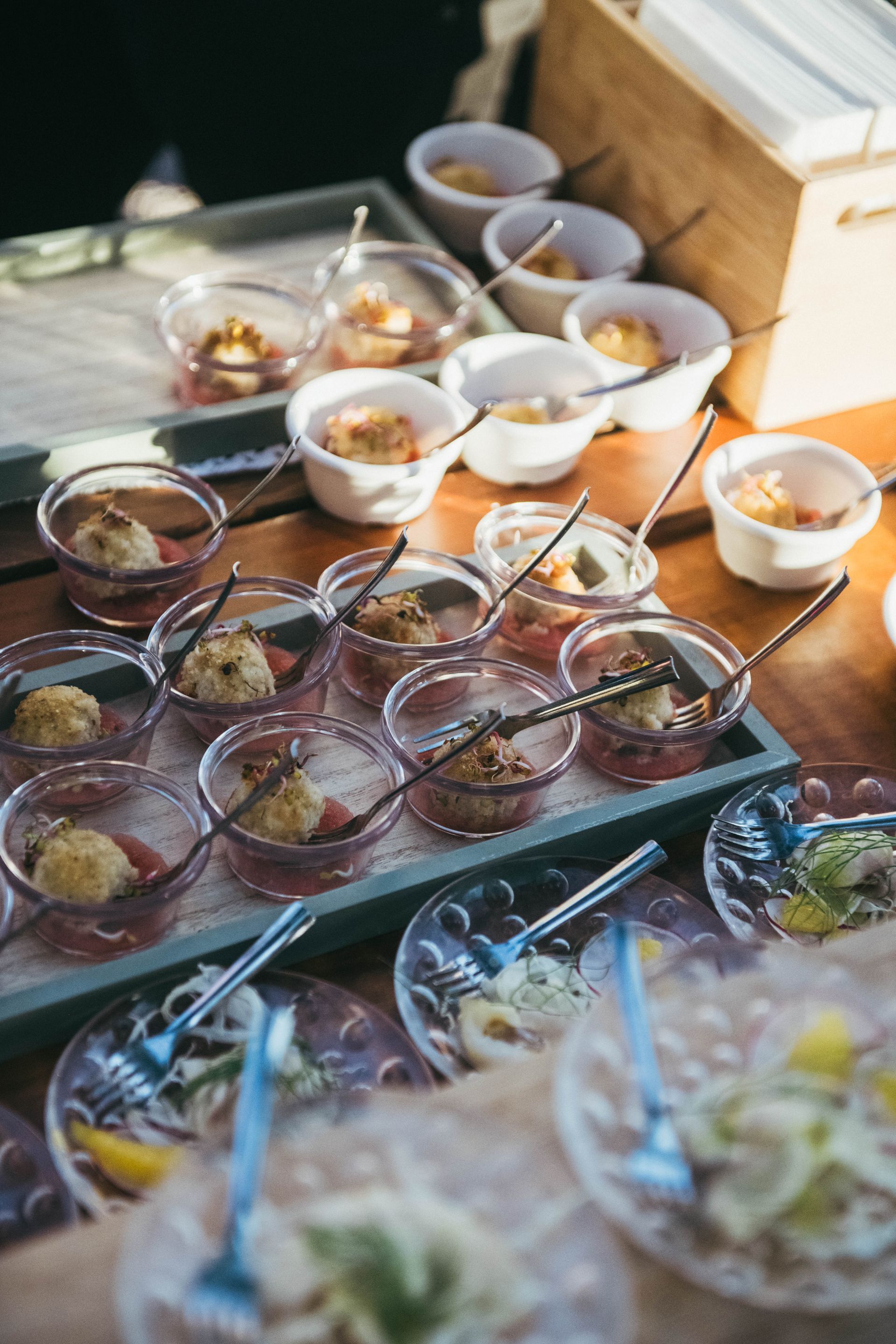 Piccole porzioni di antipasti servite con cucchiai su un tavolo di legno, con vari ingredienti in ciotole trasparenti e bianche, più insalata.