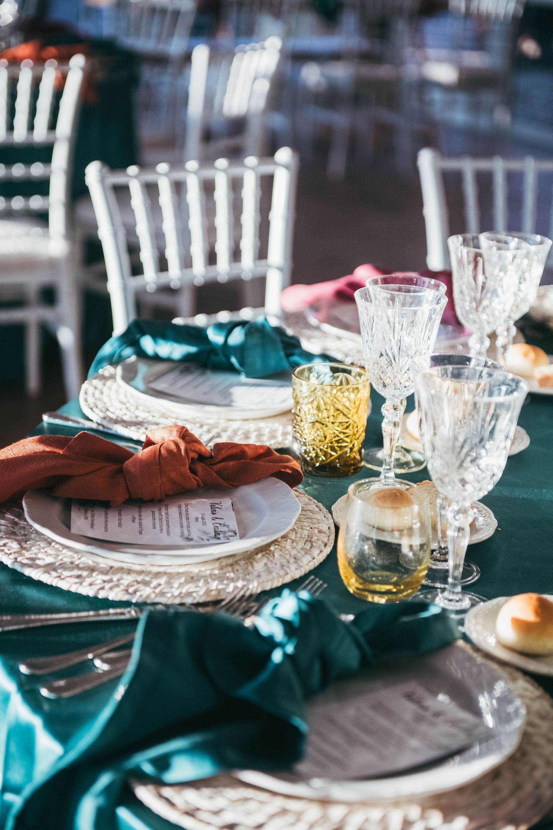 Elegante tavola apparecchiata nei toni del verde acqua e ruggine con sedie bianche. Bicchieri di cristallo, portacandele dorati e tovagliette intrecciate.