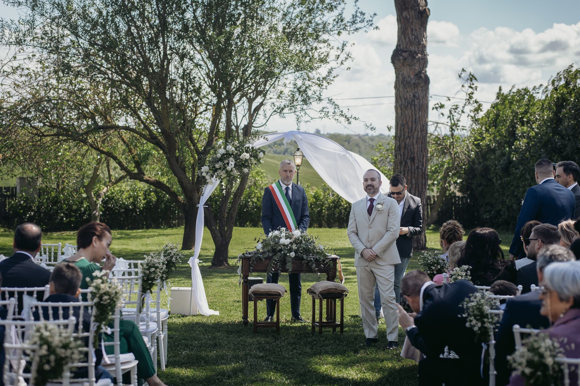 Cerimonia nuziale: lo sposo e il celebrante sono in piedi presso l'altare all'aperto, gli invitati sono seduti su sedie bianche.