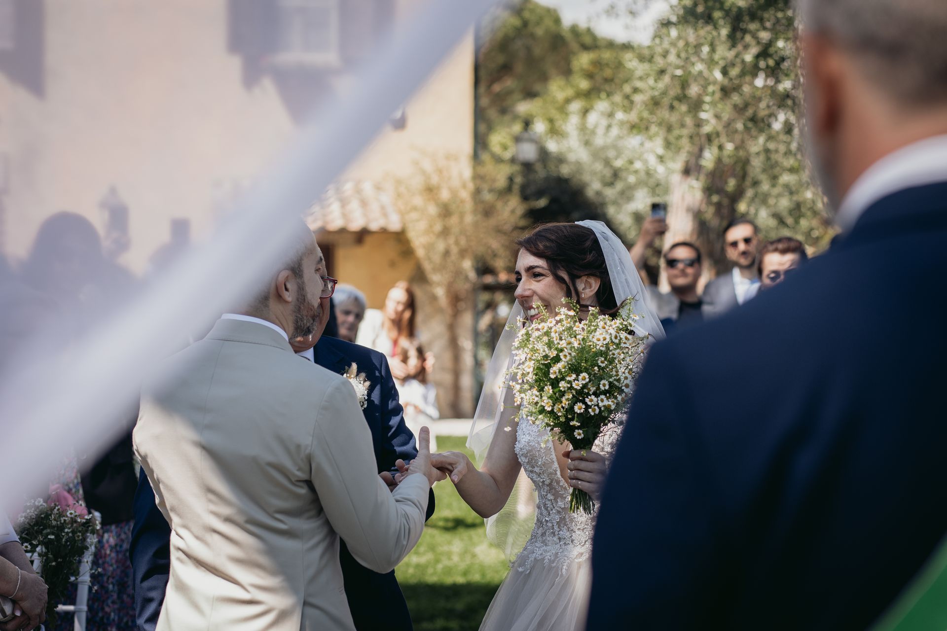 La sposa sorride e stringe la mano a un uomo durante una cerimonia nuziale all'aperto; gli invitati guardano.