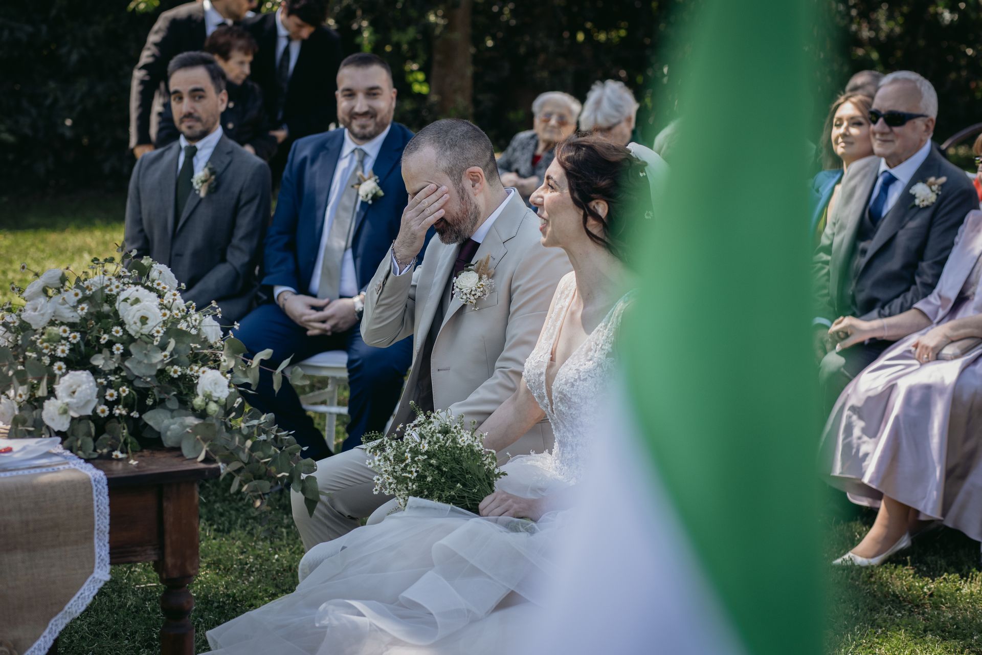 Cerimonia nuziale con lo sposo che si asciuga le lacrime, la sposa che sorride, gli invitati seduti all'aperto.