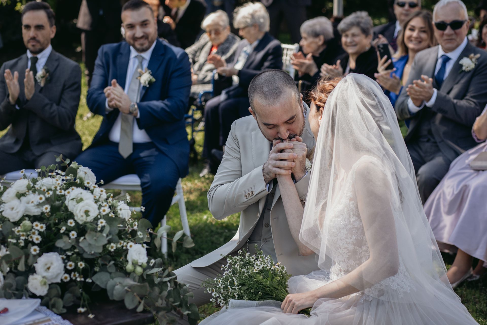 Lo sposo bacia la mano della sposa durante la cerimonia nuziale; ambientazione all'aperto, persone che applaudono.