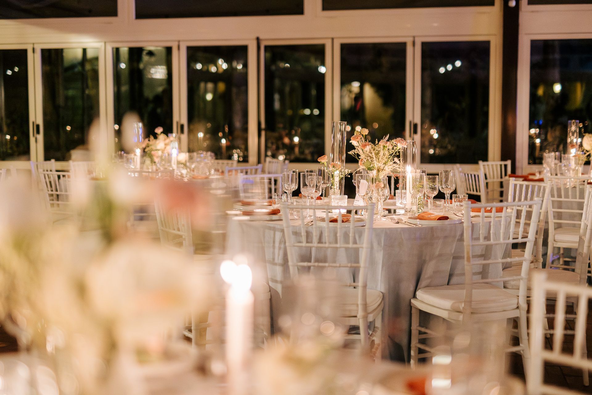 Eleganti tavoli da ricevimento apparecchiati per una festa, con fiori, candele e sedie bianche di fronte alle grandi finestre.