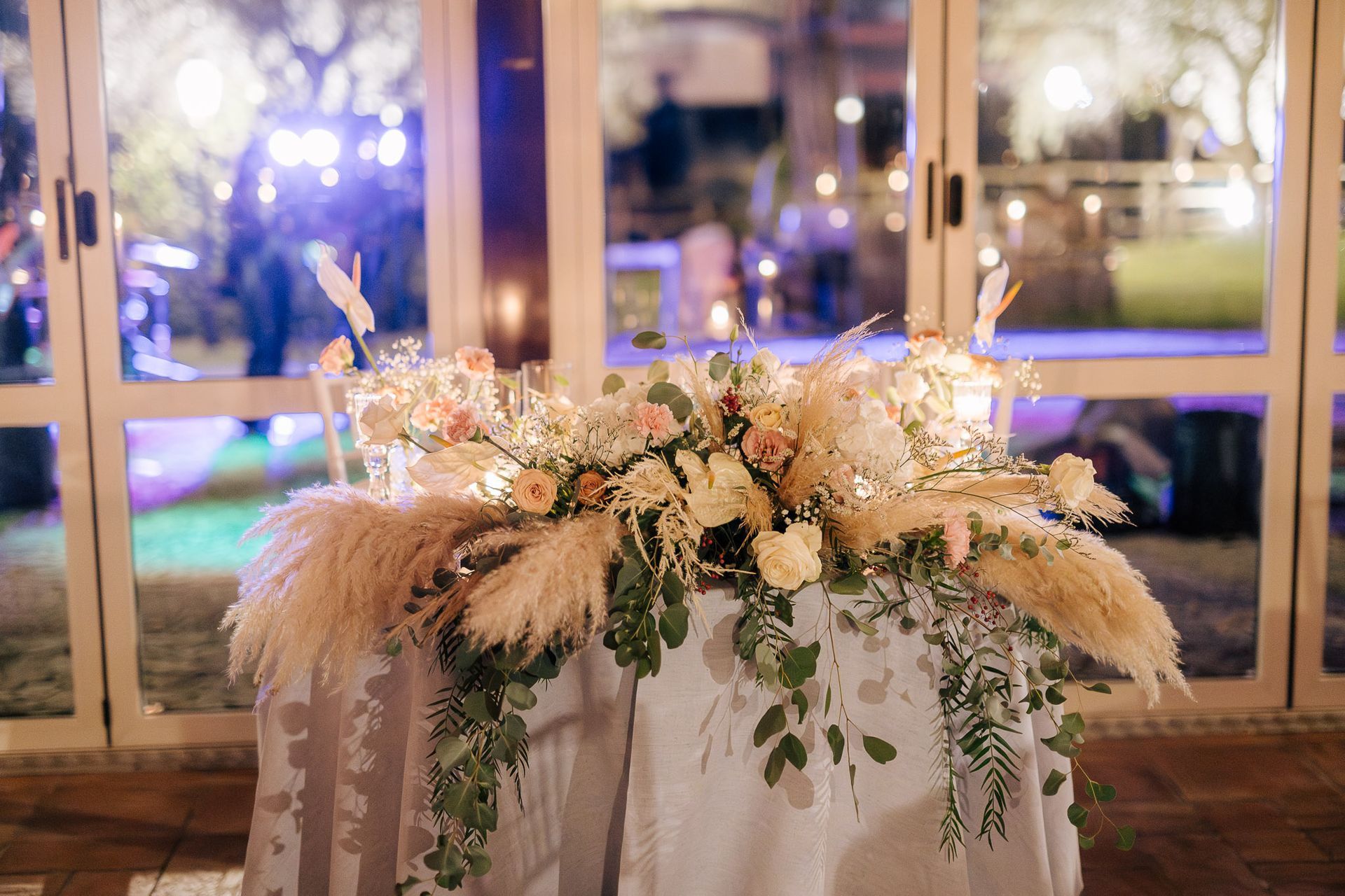 Tavolo nuziale con composizione floreale, erba della pampa e verde, sistemato davanti a porte a vetri.