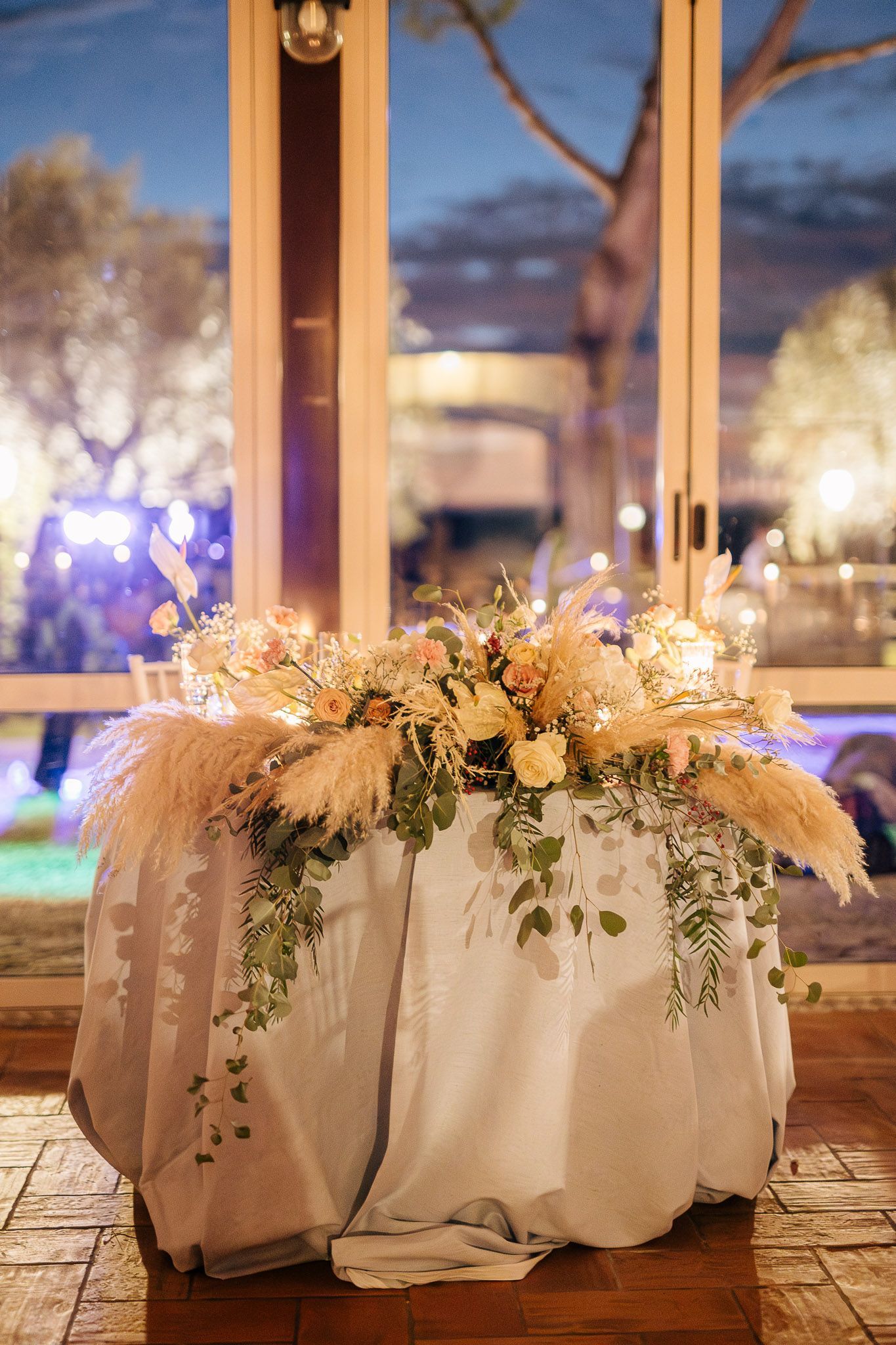 Tavolo per ricevimento di nozze con composizione floreale, erba della pampa, drappeggiato in lino bianco, con sfondo esterno.