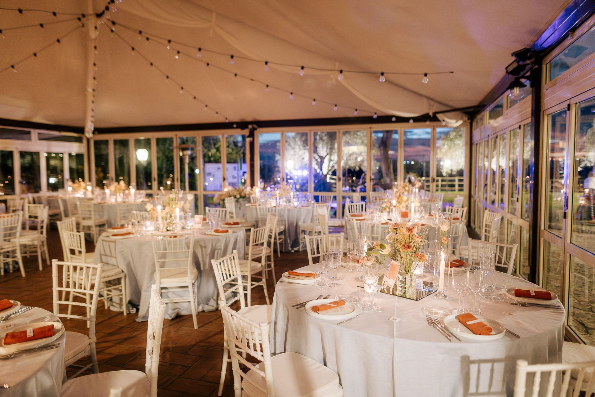 Elegante location per eventi con tavoli rotondi, sedie bianche e luci a filo; colori tenui e pareti in vetro.