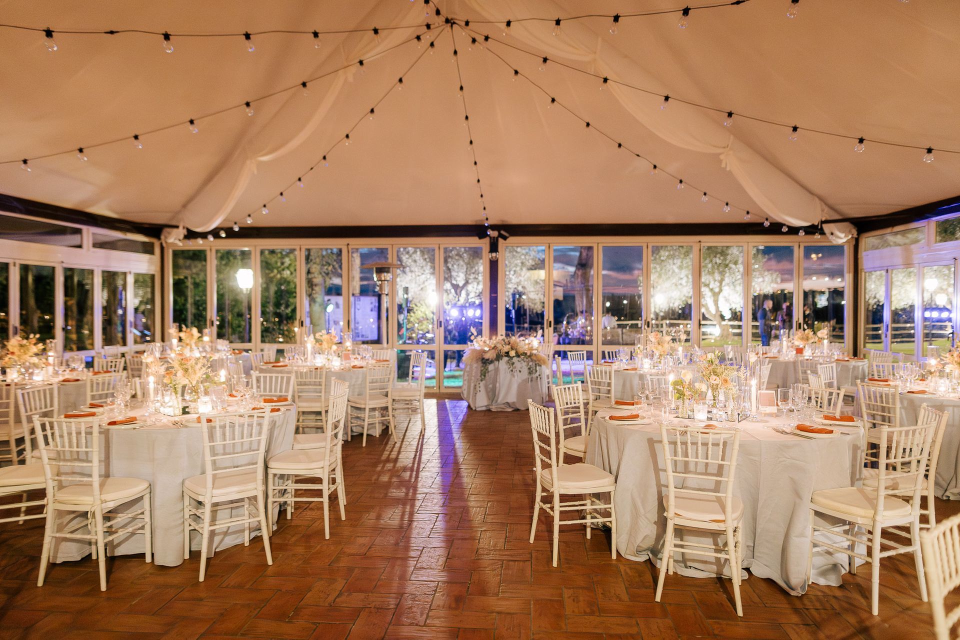 Elegante ricevimento di nozze in una tenda, con tavoli rotondi, sedie bianche e luci a filo.