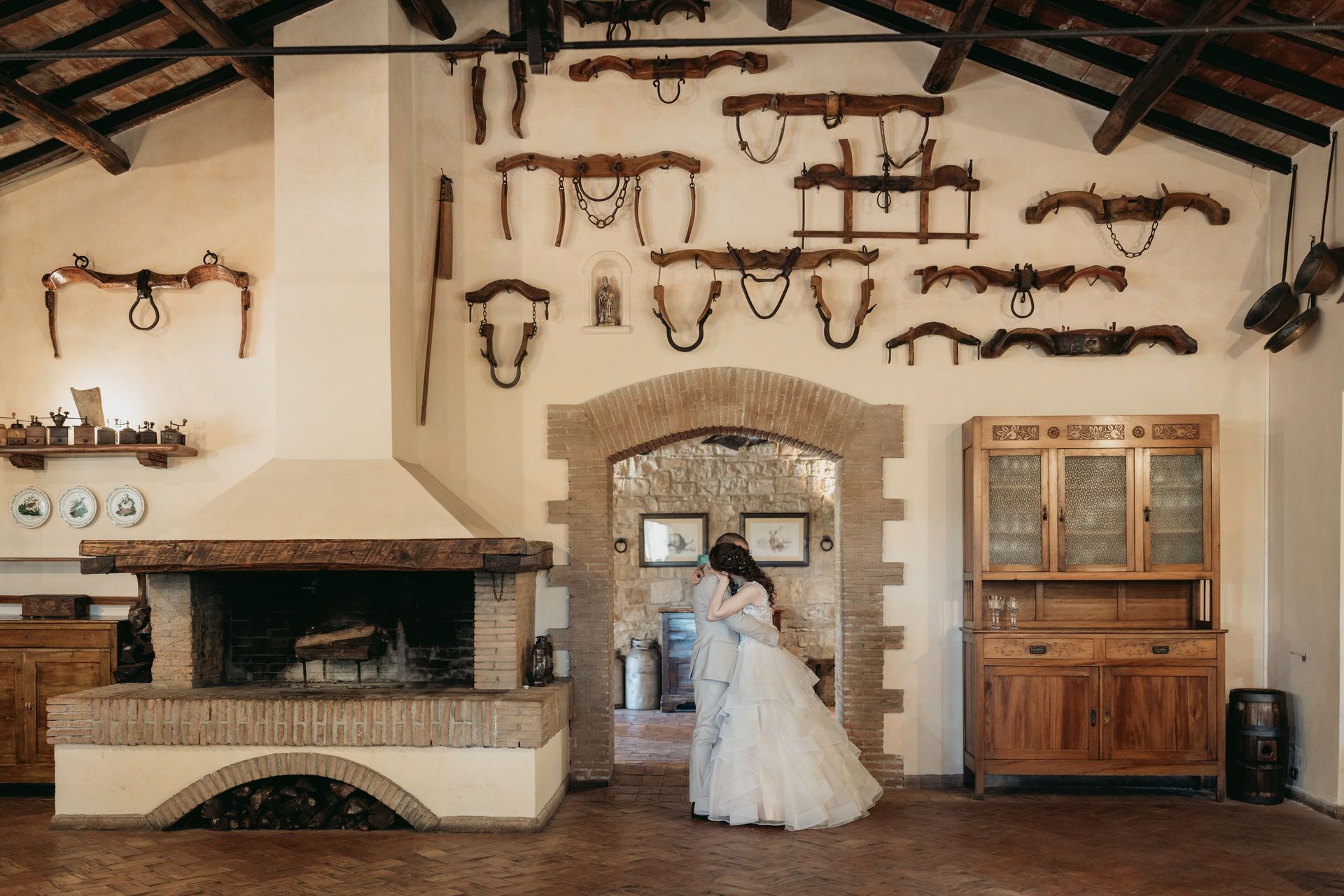 Gli sposi si abbracciano sotto un portone ad arco. Interni rustici con camino, armadi in legno e oggetti appesi alle pareti.