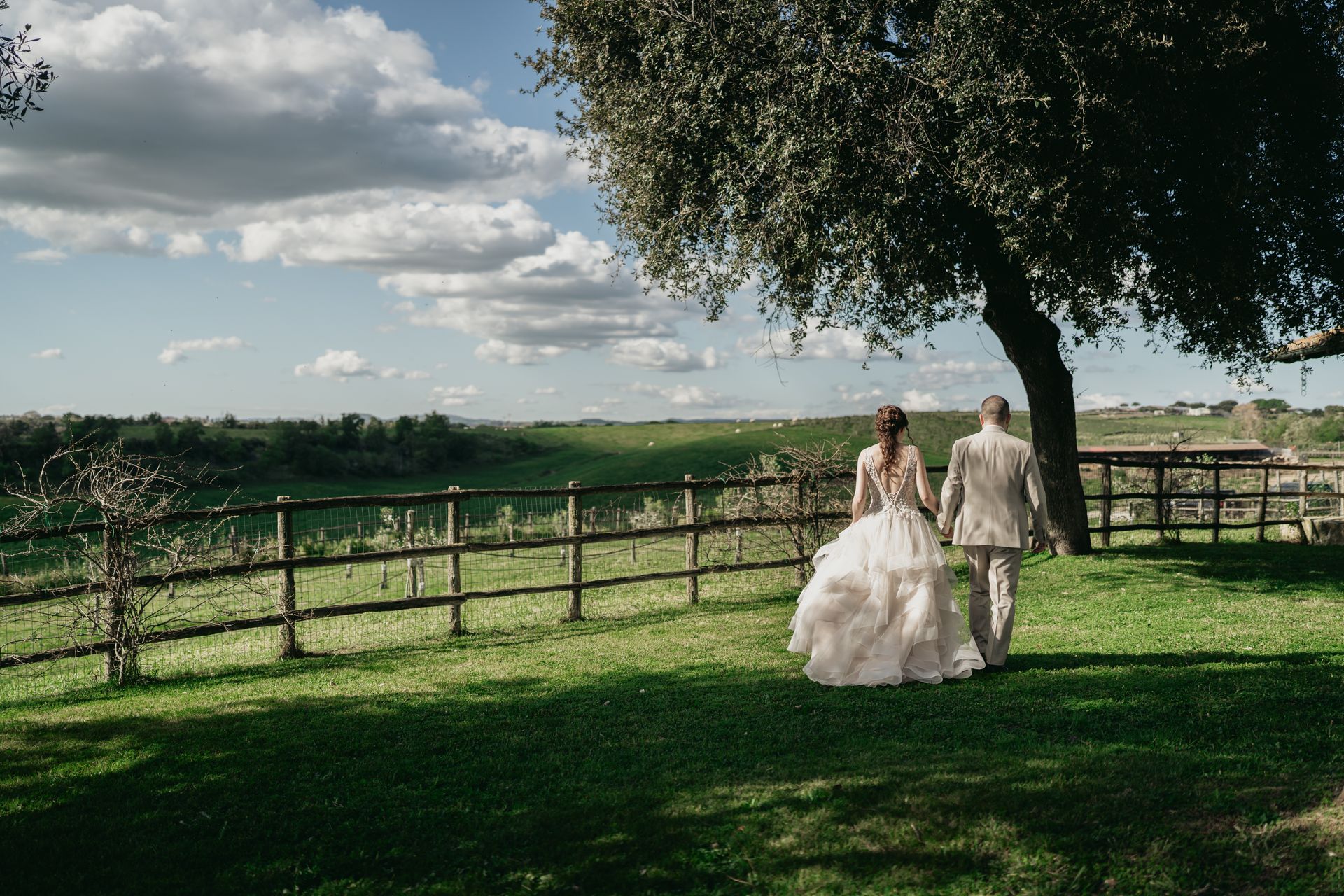 Una coppia cammina su un prato erboso, tenendosi per mano, vicino a una staccionata di legno e a un albero, sotto un cielo nuvoloso.