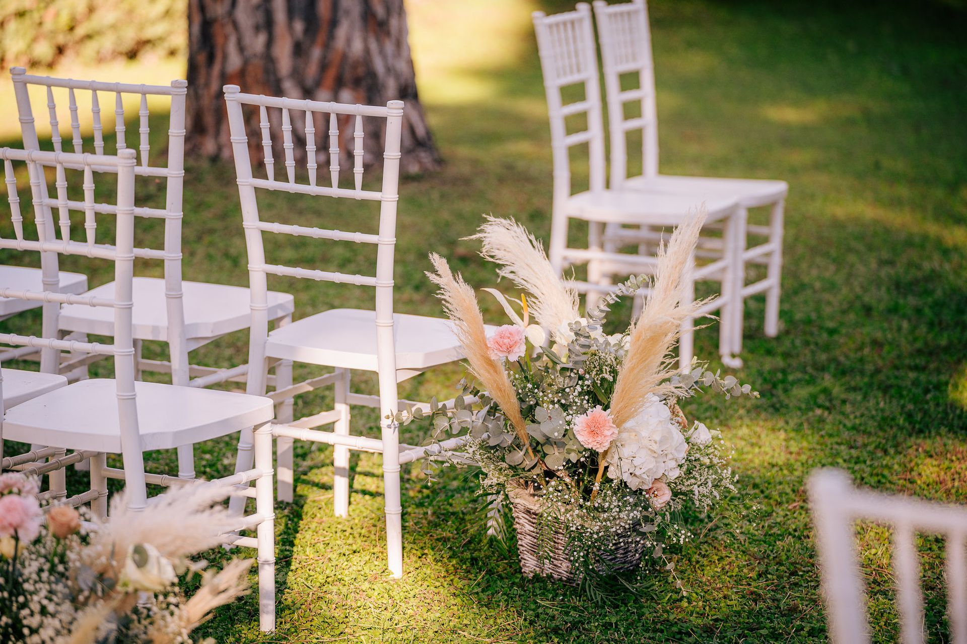 Sedie bianche allestite per una cerimonia all'aperto con composizioni floreali su un prato.