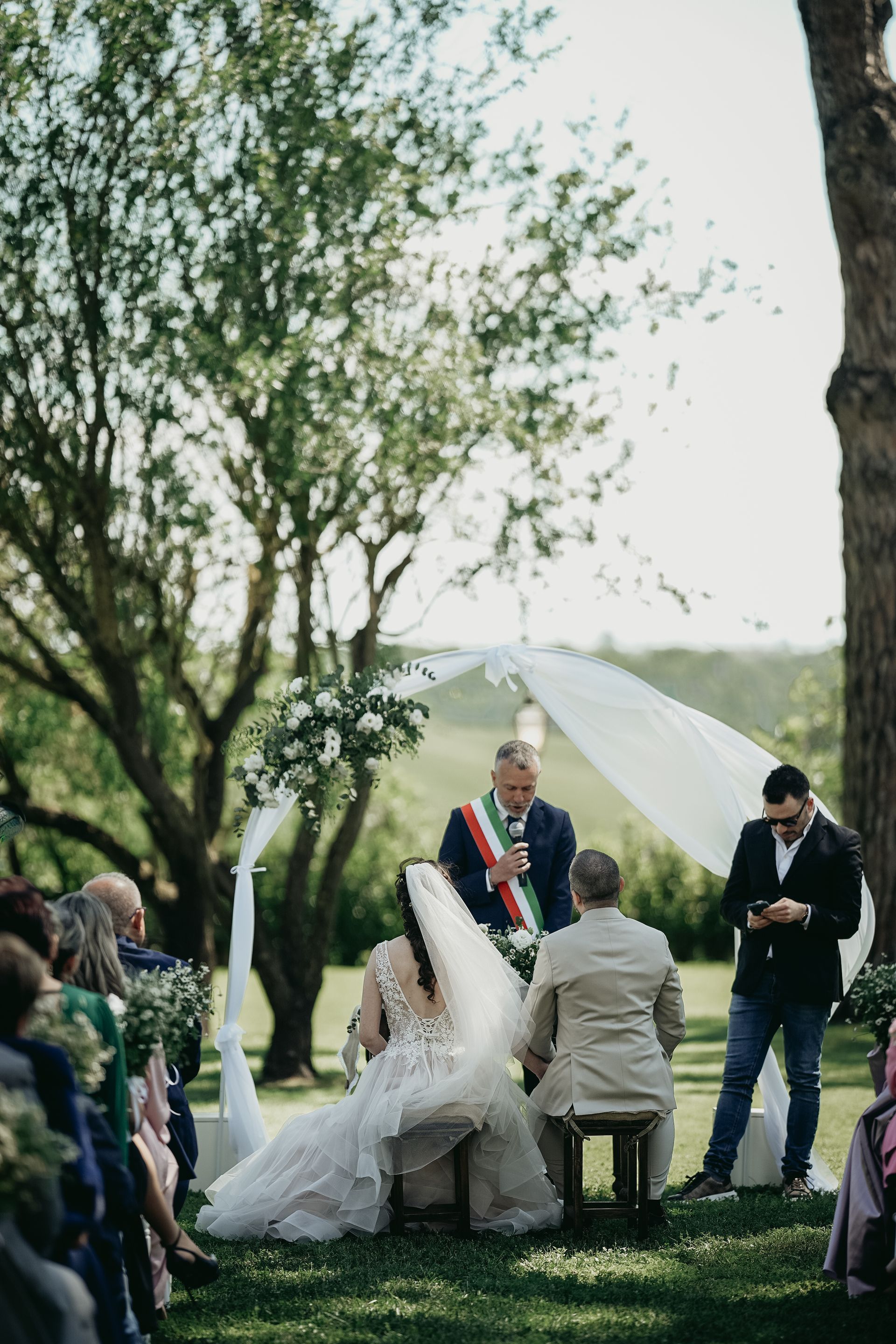 Cerimonia nuziale all'aperto: gli sposi inginocchiati, il celebrante che parla sotto l'arco drappeggiato.