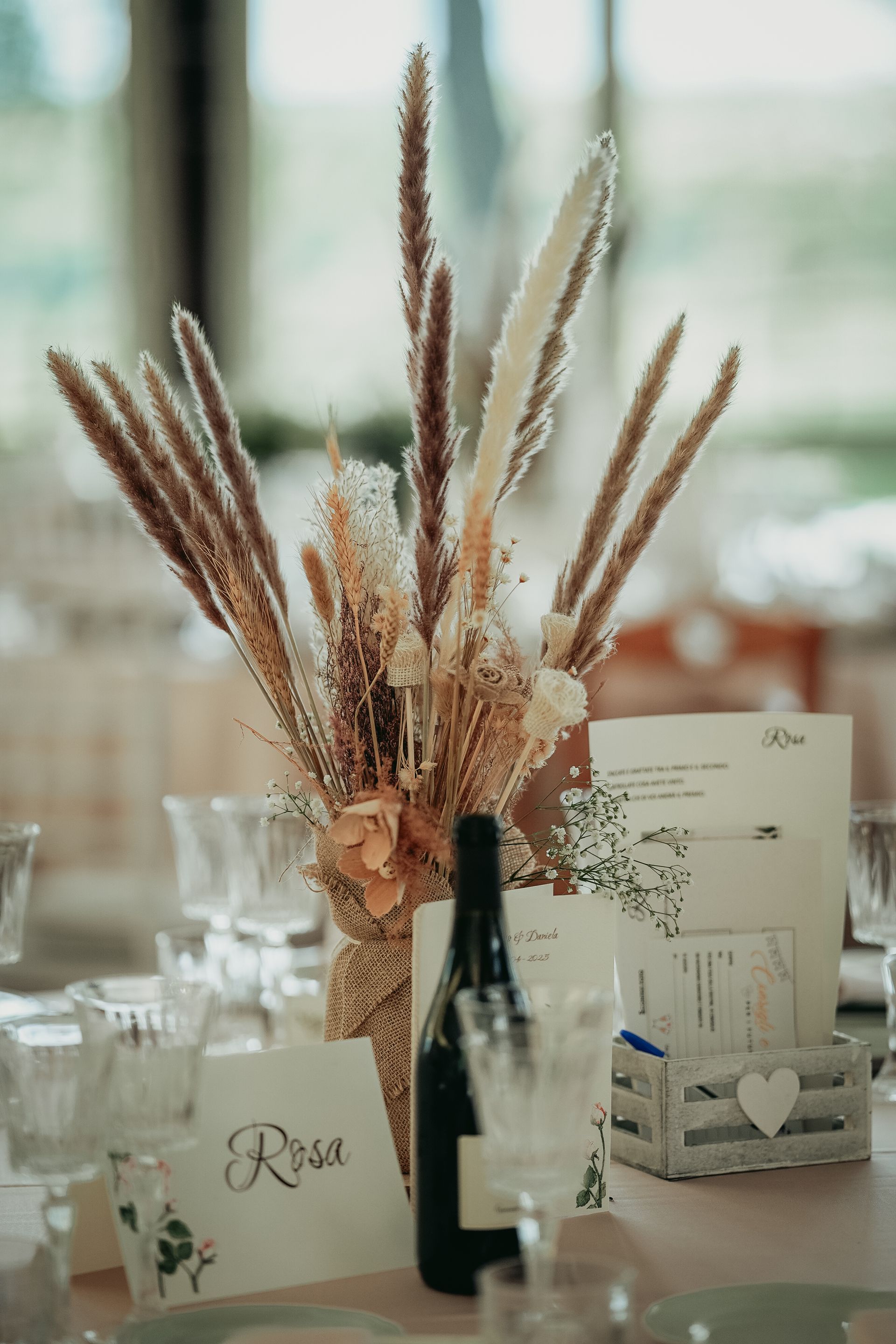 Apparecchiatura della tavola con centrotavola di fiori secchi, bottiglia di vino e segnaposto.