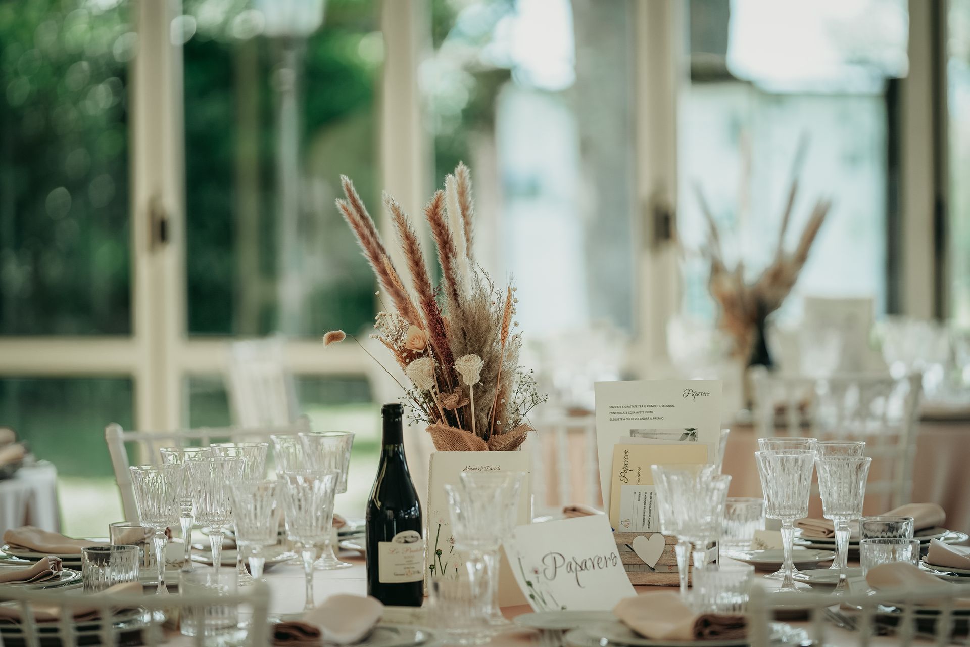 Elegante apparecchiatura da tavola con fiori secchi, bottiglia di vino e segnaposto in una stanza luminosa.