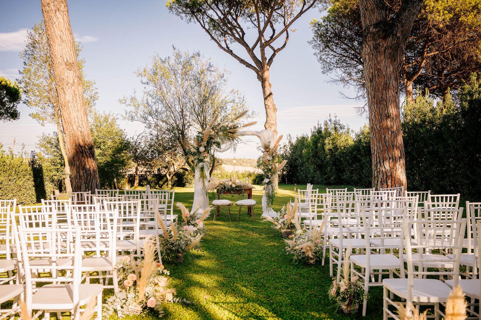 Allestimento per la cerimonia nuziale in giardino: sedie bianche, arco decorato con fiori, giornata di sole.