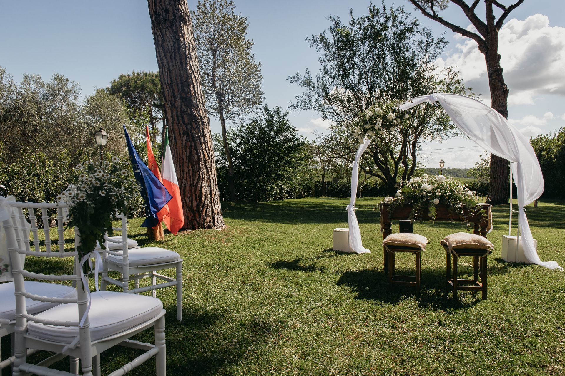 Allestimento per cerimonia nuziale all'aperto con sedie bianche, arco ricoperto di tessuto e decorazioni floreali.