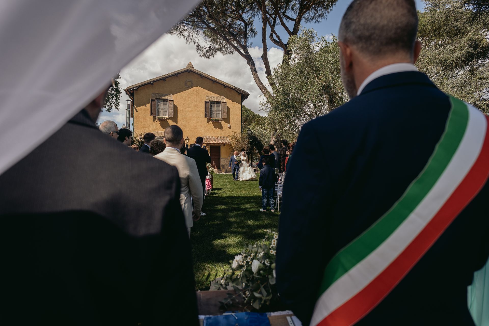 Cerimonia nuziale: gli sposi camminano verso un edificio giallo, visti da dietro gli invitati, uno dei quali indossa una fascia con i colori della bandiera italiana.