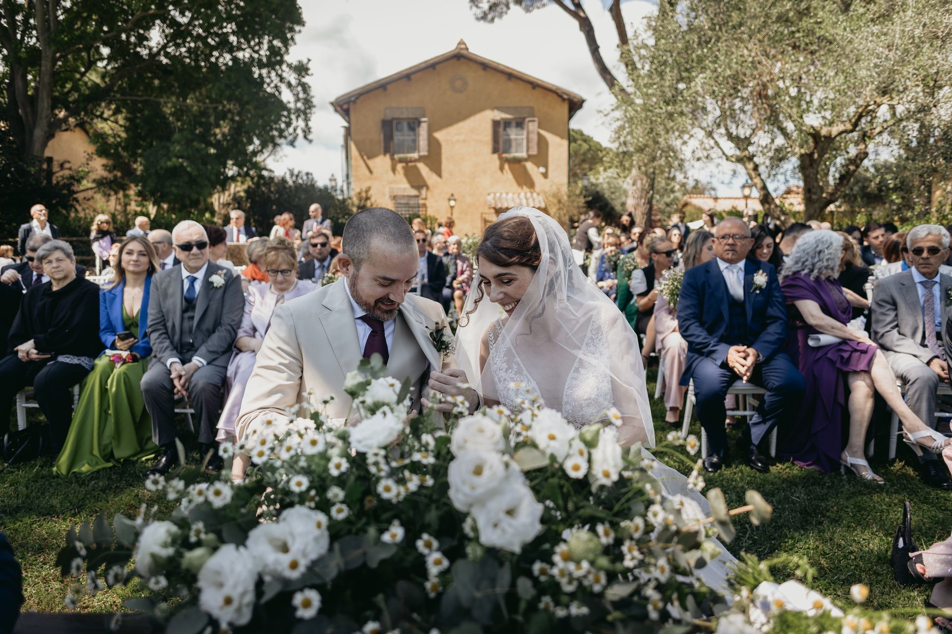 Cerimonia nuziale: coppia che si stringe le mani in una composizione floreale all'aperto, con gli ospiti seduti. Edificio beige sullo sfondo.